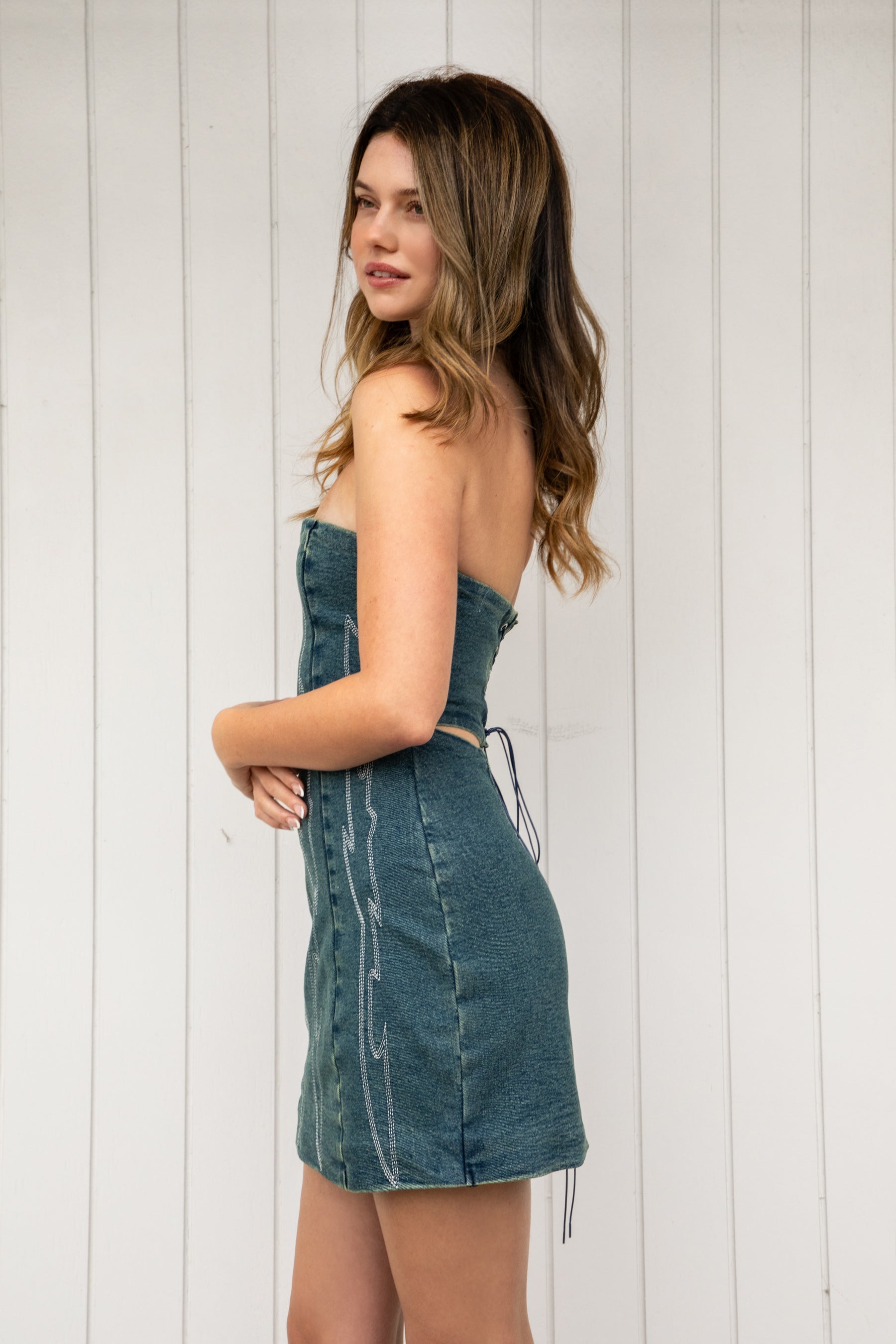 Woman wearing ARIZONA denim western stitch mini dress featuring vintage washed denim, bold western inspired contrast stitching, strapless corset style bodice, lace up back with adjustable closure, subtle back cutout, fitted waist, and a soft stretch mini length silhouette.