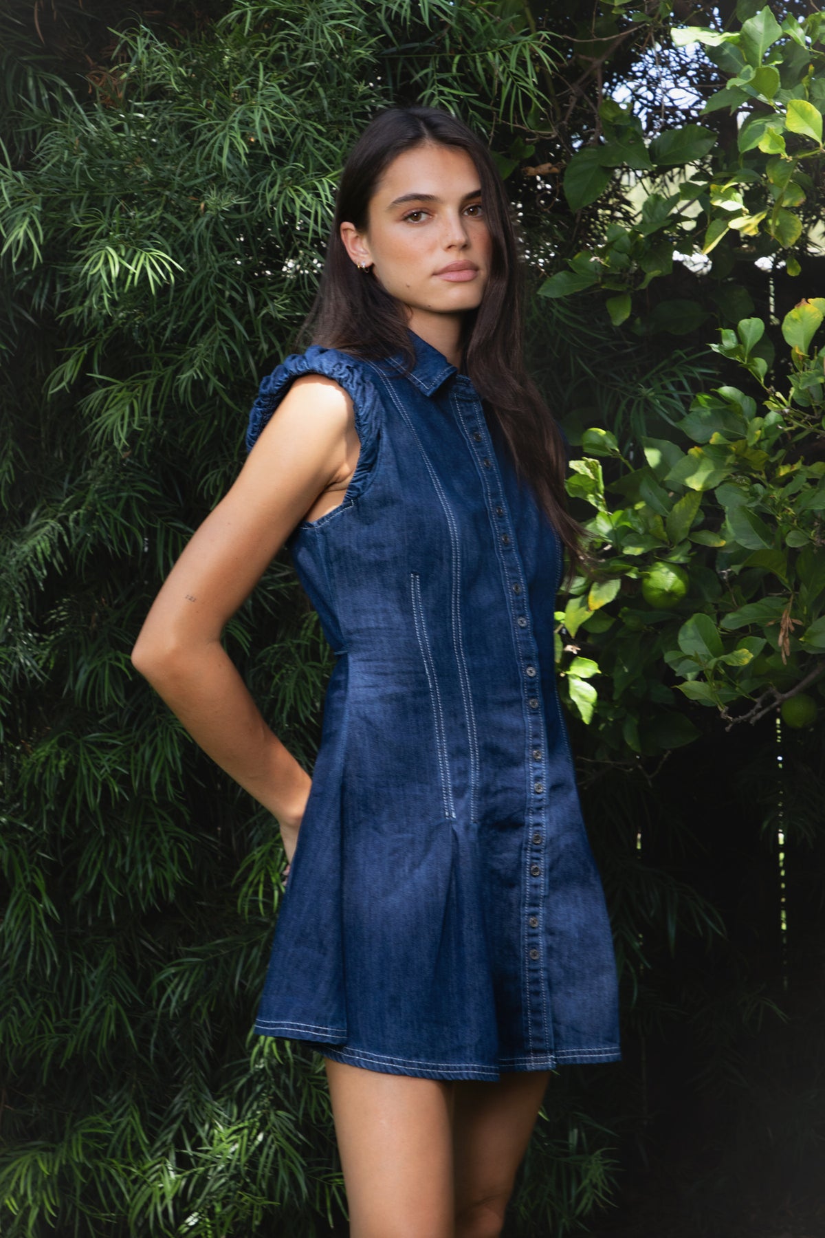 Woman wearing JOLENE ruffle sleeve button front denim mini dress featuring classic cotton blend denim fabric with a subtle washed finish, collared neckline with full front button closure, gathered ruffle cap sleeves, fitted bodice with princess seam detailing, adjustable back tie for a defined waistline, flared mini skirt with pleated details, and contrast stitching throughout.