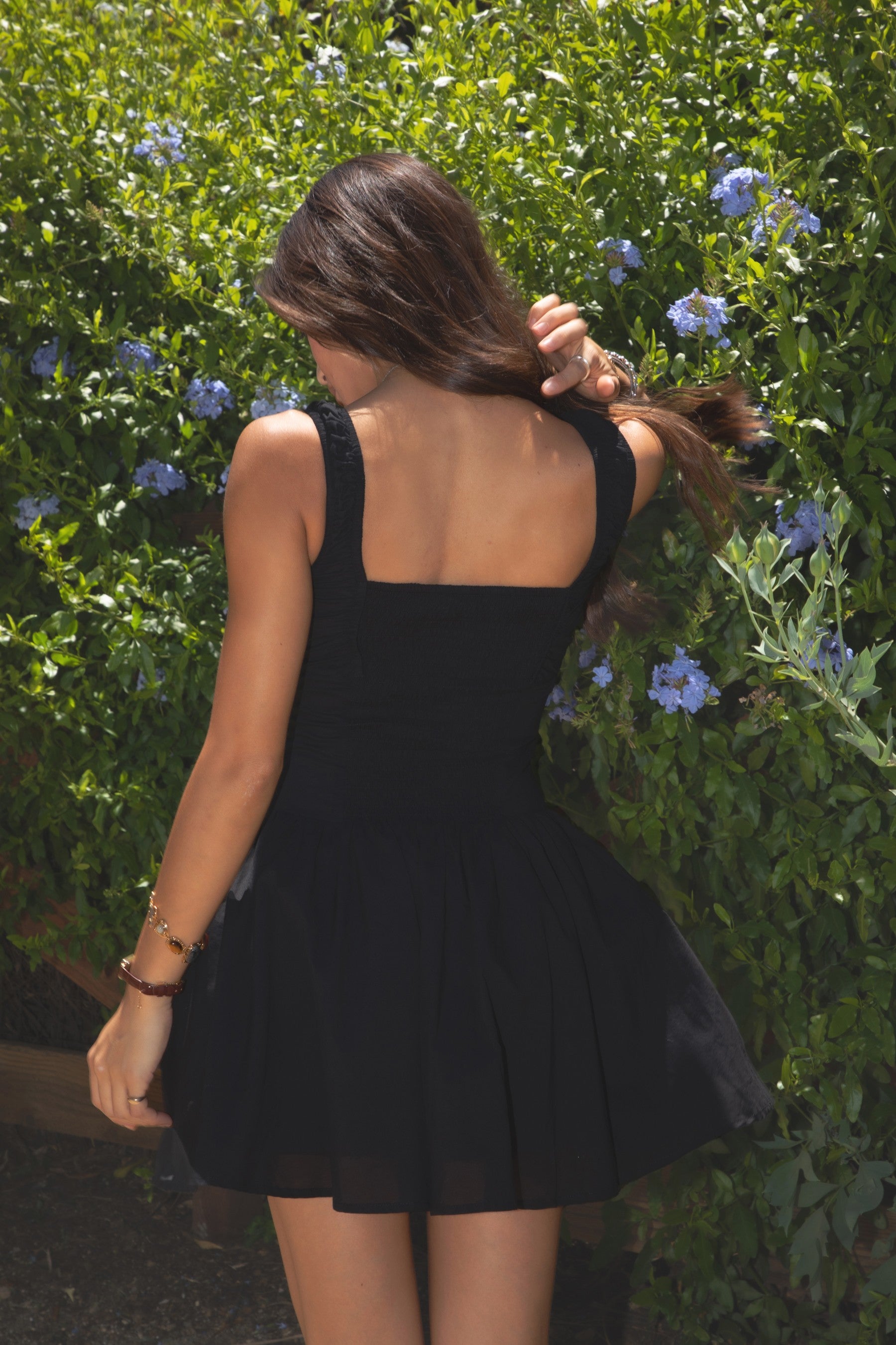 Woman wearing TIC TOC JENAH Poplin Ruffle Mini Dress in black with square neckline, tie-front bust, ruffle straps, and faux button placket standing outdoors with greenery in the background. 