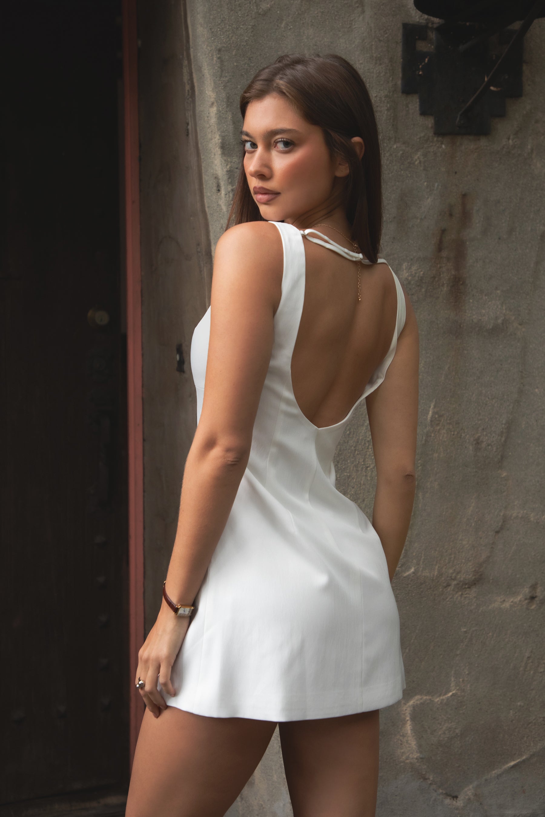 Woman wearing TIC TOC DH31914-ALEINA ALEINA mini dress made from sleek stretch fabric, featuring a clean high boat neckline, minimal sleeveless silhouette, subtle bust shaping, modern micro mini length, and open back detail with an adjustable back strap, styled with heels and a small shoulder bag for a summer night out look.