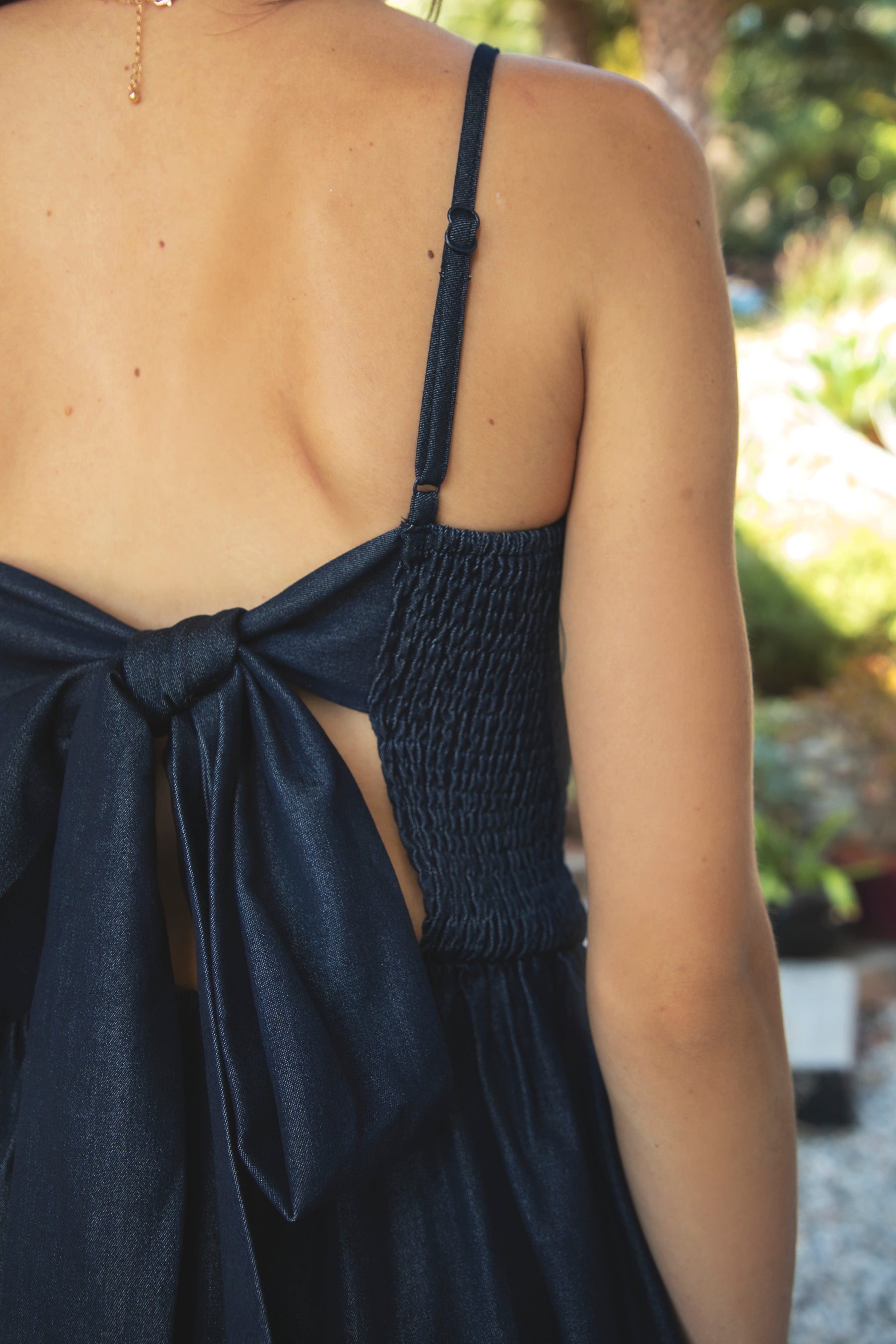Woman wearing MAYREN smocked bow back denim Tencel maxi dress featuring soft denim Tencel fabric, comfortable stretch, straight neckline, adjustable spaghetti straps, smocked bodice, statement open back with oversized self tie bow detail, side seam pockets, and flowing maxi length skirt.