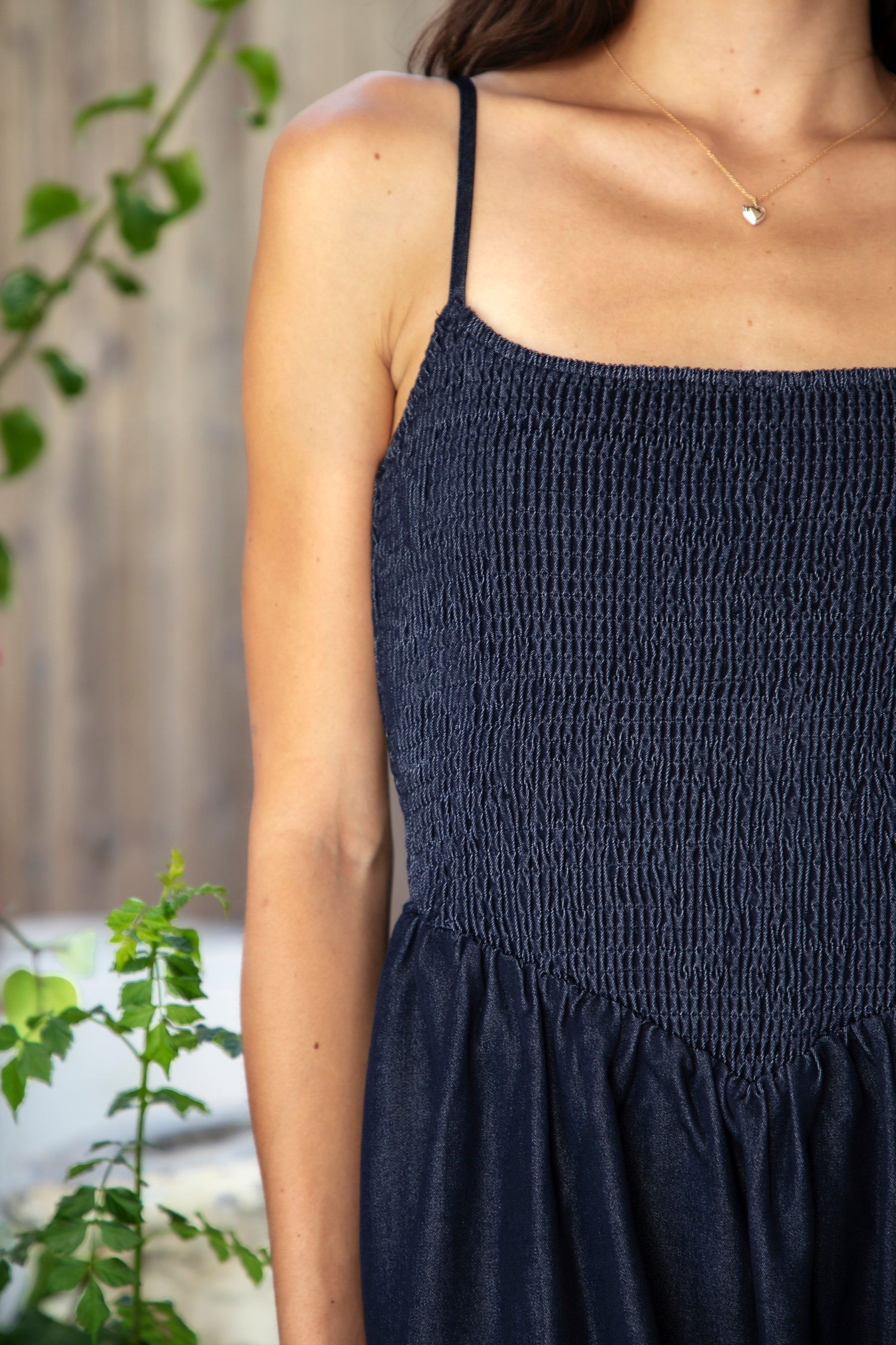 Woman wearing MAYREN smocked bow back denim Tencel maxi dress featuring soft denim Tencel fabric, comfortable stretch, straight neckline, adjustable spaghetti straps, smocked bodice, statement open back with oversized self tie bow detail, side seam pockets, and flowing maxi length skirt.
