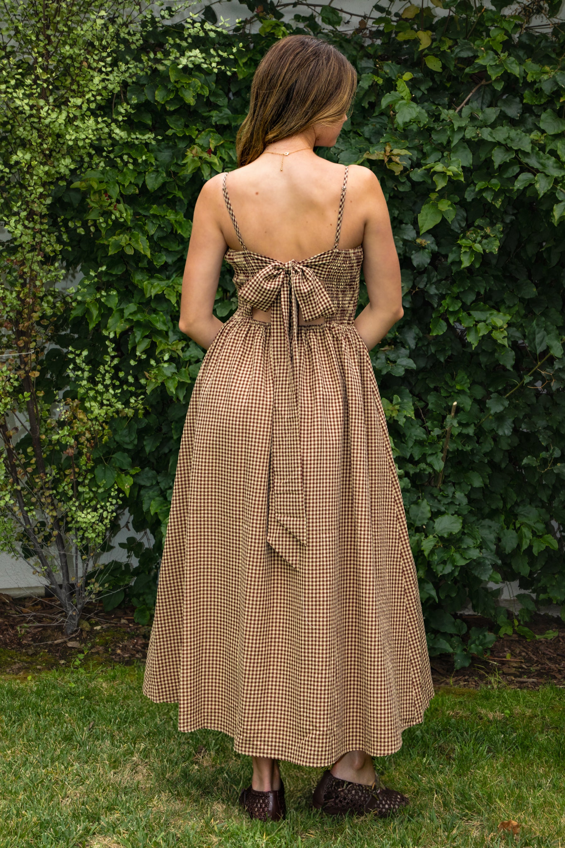 Woman wearing MAYREN gingham smocked midi dress featuring a classic gingham check pattern, fully smocked bodice, adjustable spaghetti straps, square neckline, open back with adjustable tie detail, subtle cutout accent, side seam pockets, and a flowing gathered midi skirt.