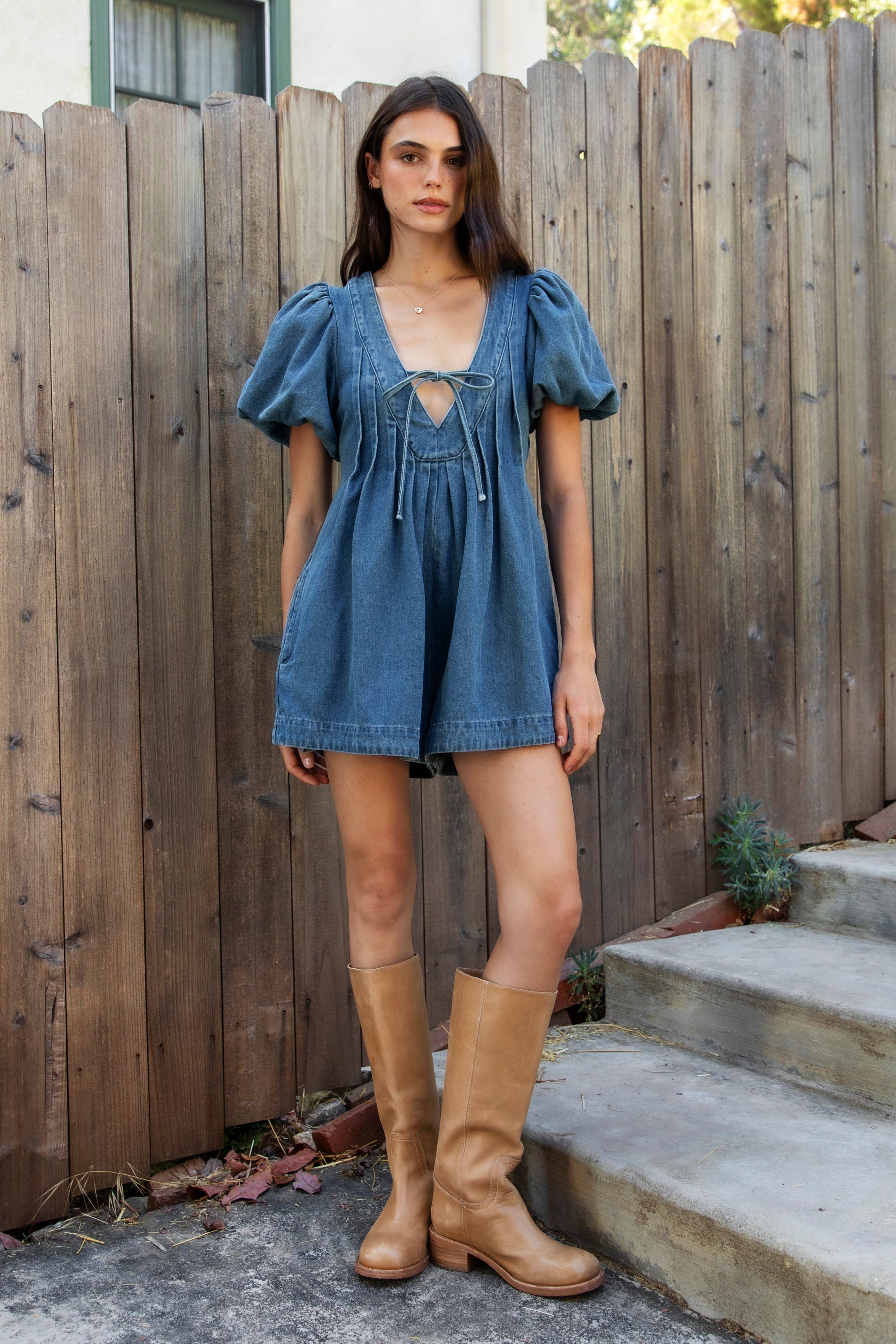 Woman wearing DELANEY soft denim puff sleeve denim romper featuring soft denim fabric, comfortable structured feel, deep V neckline, adjustable front tie detail, statement puff sleeves with gathered volume, pleated bodice, relaxed fit silhouette, and hidden side zipper closure.