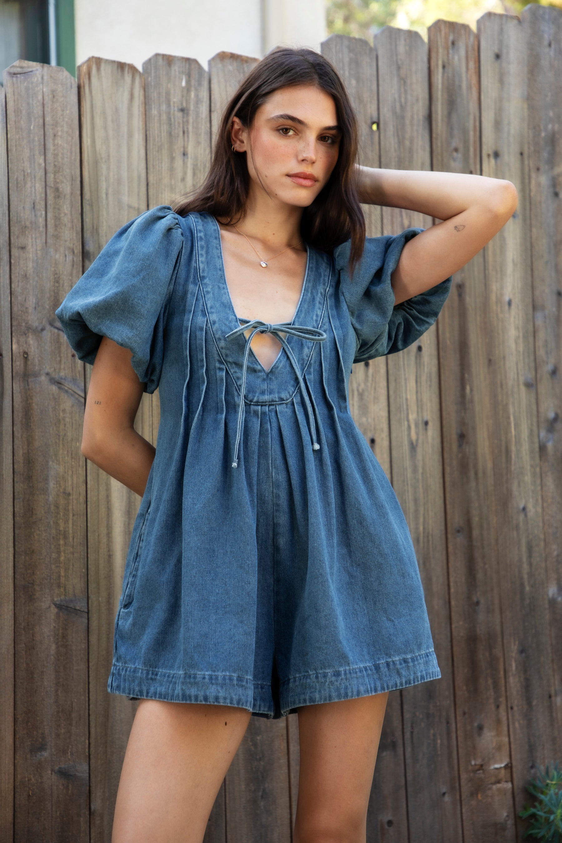 Woman wearing DELANEY soft denim puff sleeve denim romper featuring soft denim fabric, comfortable structured feel, deep V neckline, adjustable front tie detail, statement puff sleeves with gathered volume, pleated bodice, relaxed fit silhouette, and hidden side zipper closure.
