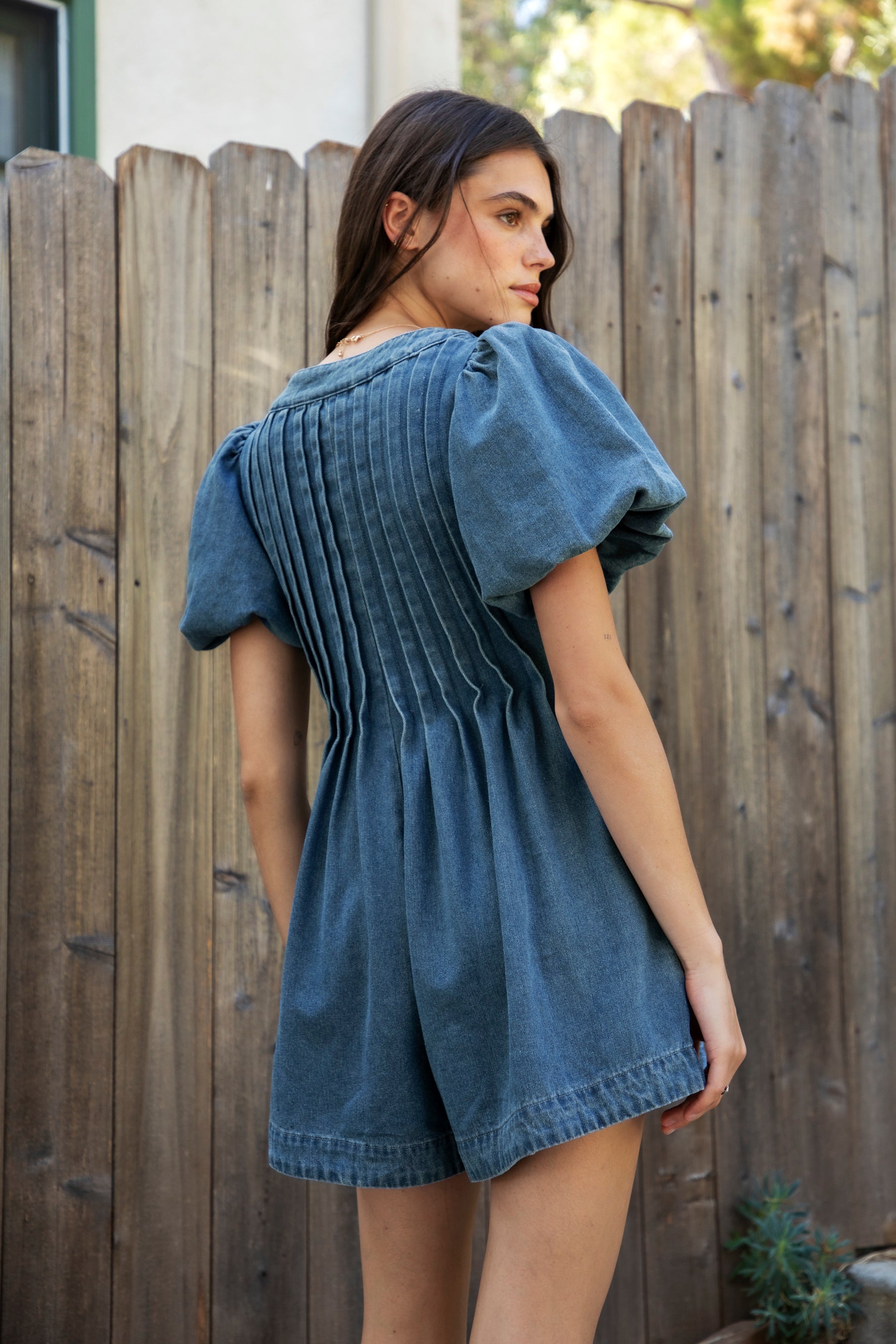 Woman wearing DELANEY soft denim puff sleeve denim romper featuring soft denim fabric, comfortable structured feel, deep V neckline, adjustable front tie detail, statement puff sleeves with gathered volume, pleated bodice, relaxed fit silhouette, and hidden side zipper closure.