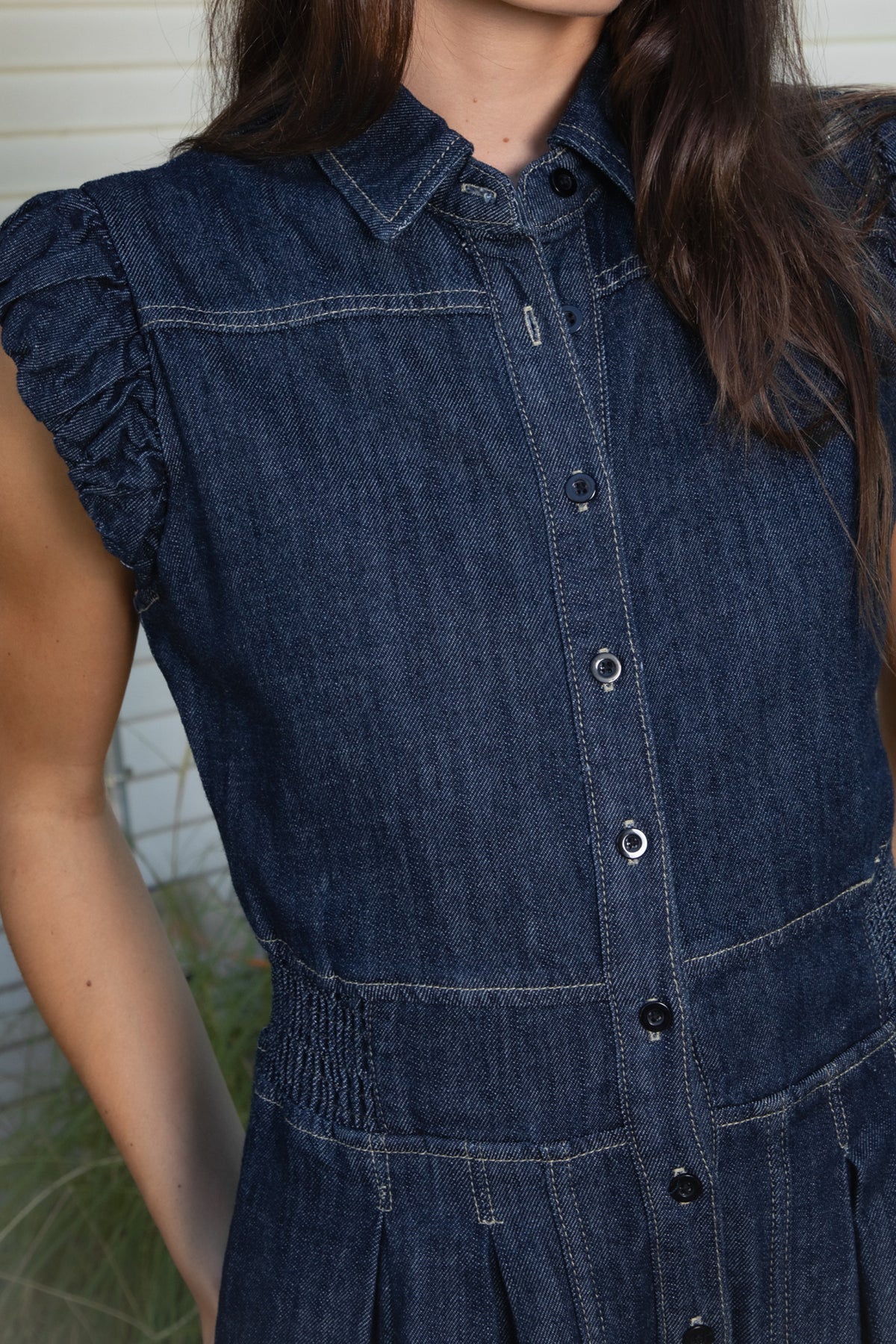 Woman wearing SHILOW ruffle sleeve wide leg denim jumpsuit featuring classic dark blue cotton blend denim fabric, collared neckline with front button closure, feminine gathered ruffle cap sleeves, smocked elastic waistband, front pleated detailing, relaxed balloon leg silhouette with cropped length, adjustable drawstring details at the leg hems, functional side pockets, back patch pockets, and contrast stitching throughout.