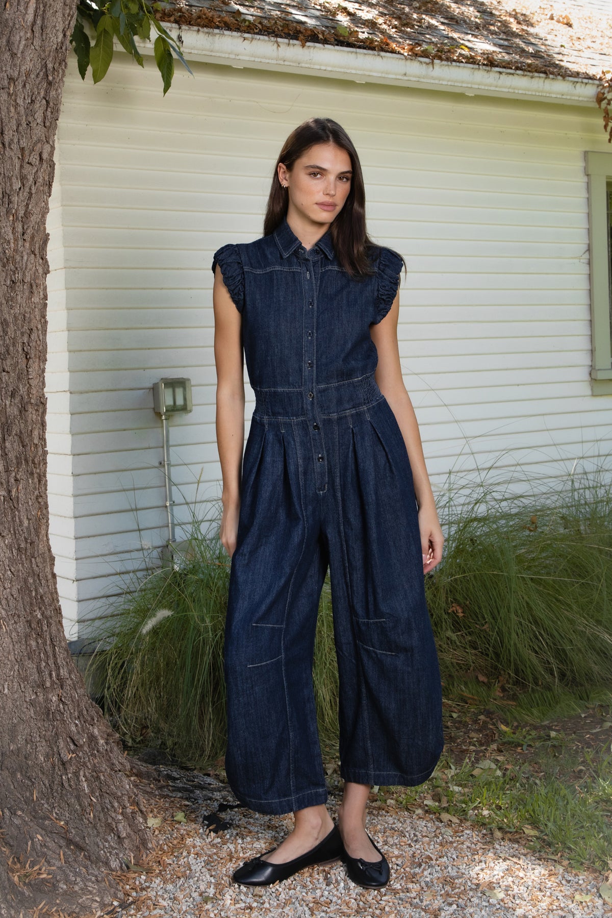 Woman wearing SHILOW ruffle sleeve wide leg denim jumpsuit featuring classic dark blue cotton blend denim fabric, collared neckline with front button closure, feminine gathered ruffle cap sleeves, smocked elastic waistband, front pleated detailing, relaxed balloon leg silhouette with cropped length, adjustable drawstring details at the leg hems, functional side pockets, back patch pockets, and contrast stitching throughout.