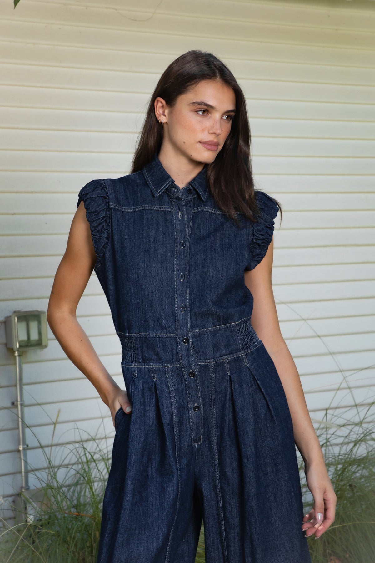 Woman wearing SHILOW ruffle sleeve wide leg denim jumpsuit featuring classic dark blue cotton blend denim fabric, collared neckline with front button closure, feminine gathered ruffle cap sleeves, smocked elastic waistband, front pleated detailing, relaxed balloon leg silhouette with cropped length, adjustable drawstring details at the leg hems, functional side pockets, back patch pockets, and contrast stitching throughout.