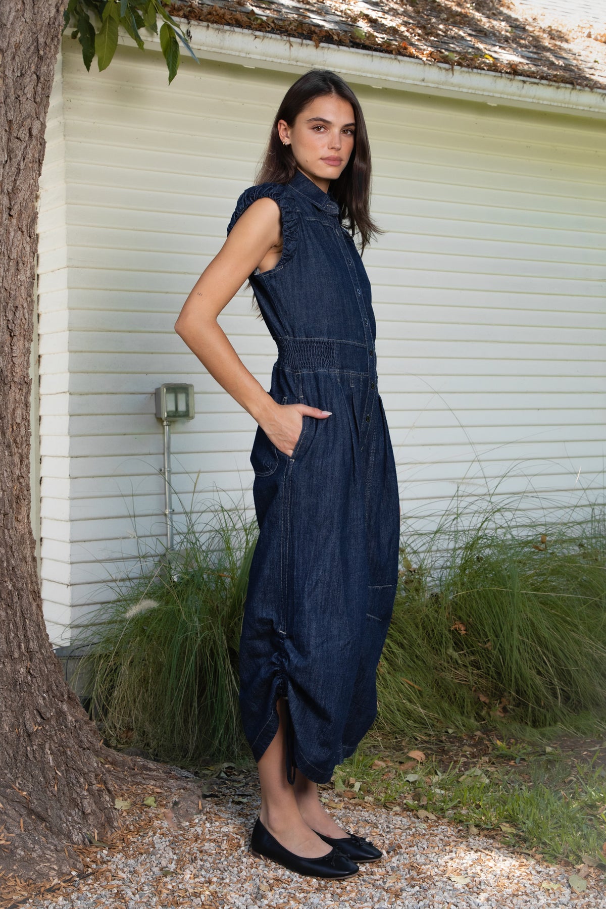 Woman wearing SHILOW ruffle sleeve wide leg denim jumpsuit featuring classic dark blue cotton blend denim fabric, collared neckline with front button closure, feminine gathered ruffle cap sleeves, smocked elastic waistband, front pleated detailing, relaxed balloon leg silhouette with cropped length, adjustable drawstring details at the leg hems, functional side pockets, back patch pockets, and contrast stitching throughout.