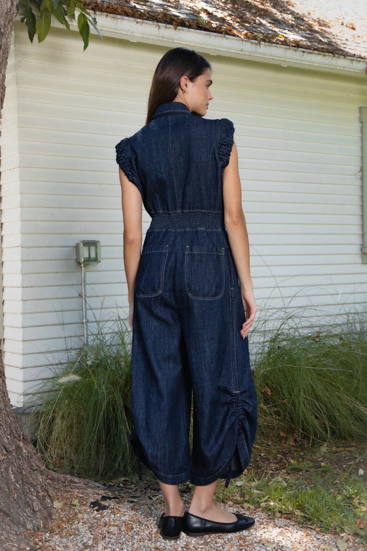 Woman wearing SHILOW ruffle sleeve wide leg denim jumpsuit featuring classic dark blue cotton blend denim fabric, collared neckline with front button closure, feminine gathered ruffle cap sleeves, smocked elastic waistband, front pleated detailing, relaxed balloon leg silhouette with cropped length, adjustable drawstring details at the leg hems, functional side pockets, back patch pockets, and contrast stitching throughout.