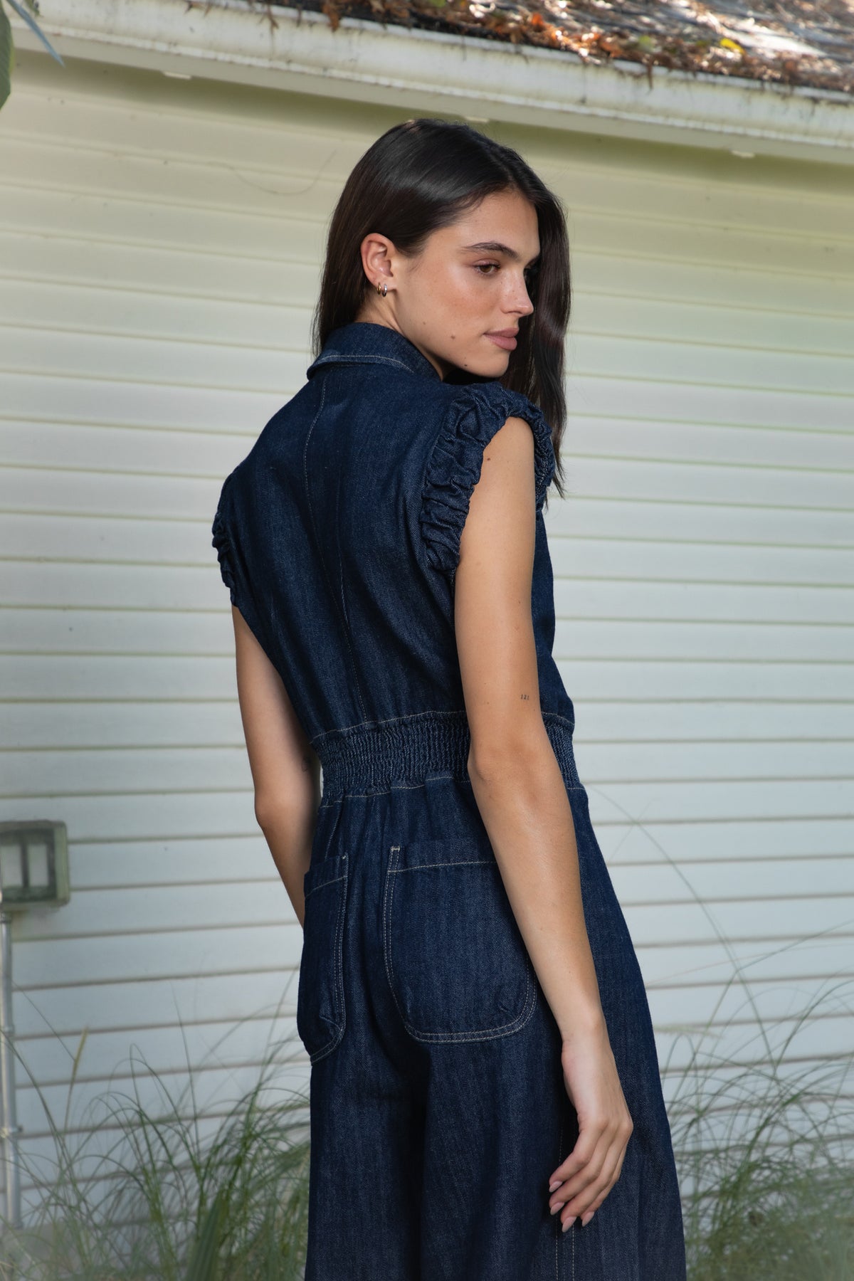 Woman wearing SHILOW ruffle sleeve wide leg denim jumpsuit featuring classic dark blue cotton blend denim fabric, collared neckline with front button closure, feminine gathered ruffle cap sleeves, smocked elastic waistband, front pleated detailing, relaxed balloon leg silhouette with cropped length, adjustable drawstring details at the leg hems, functional side pockets, back patch pockets, and contrast stitching throughout.
