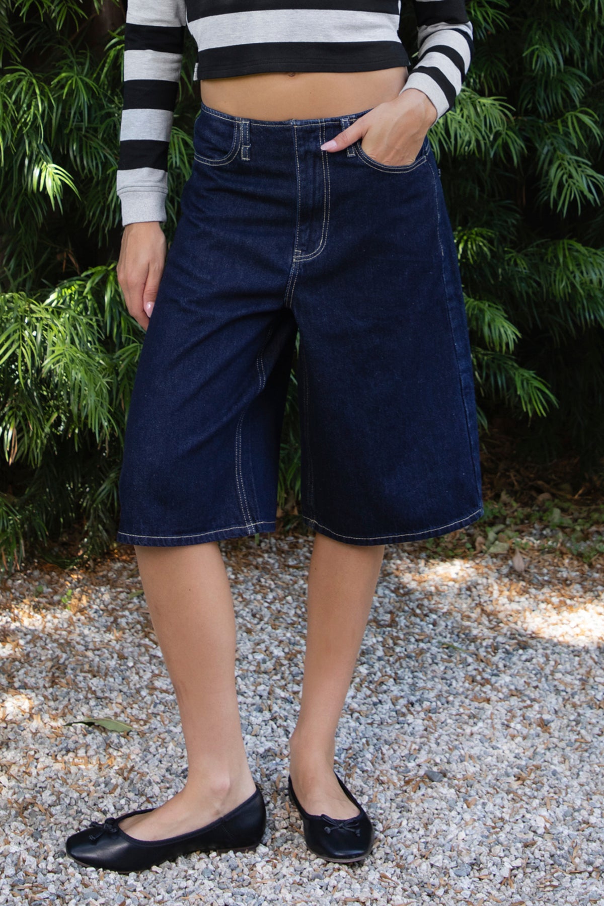 Woman wearing PIPER wide leg denim bermuda shorts featuring classic dark blue cotton blend denim fabric, mid rise waistline with belt loops, relaxed fit through the hips and thighs, wide leg knee length silhouette, front button and zipper fly closure, classic five pocket design, contrast stitching throughout, and a straight clean finished hem.