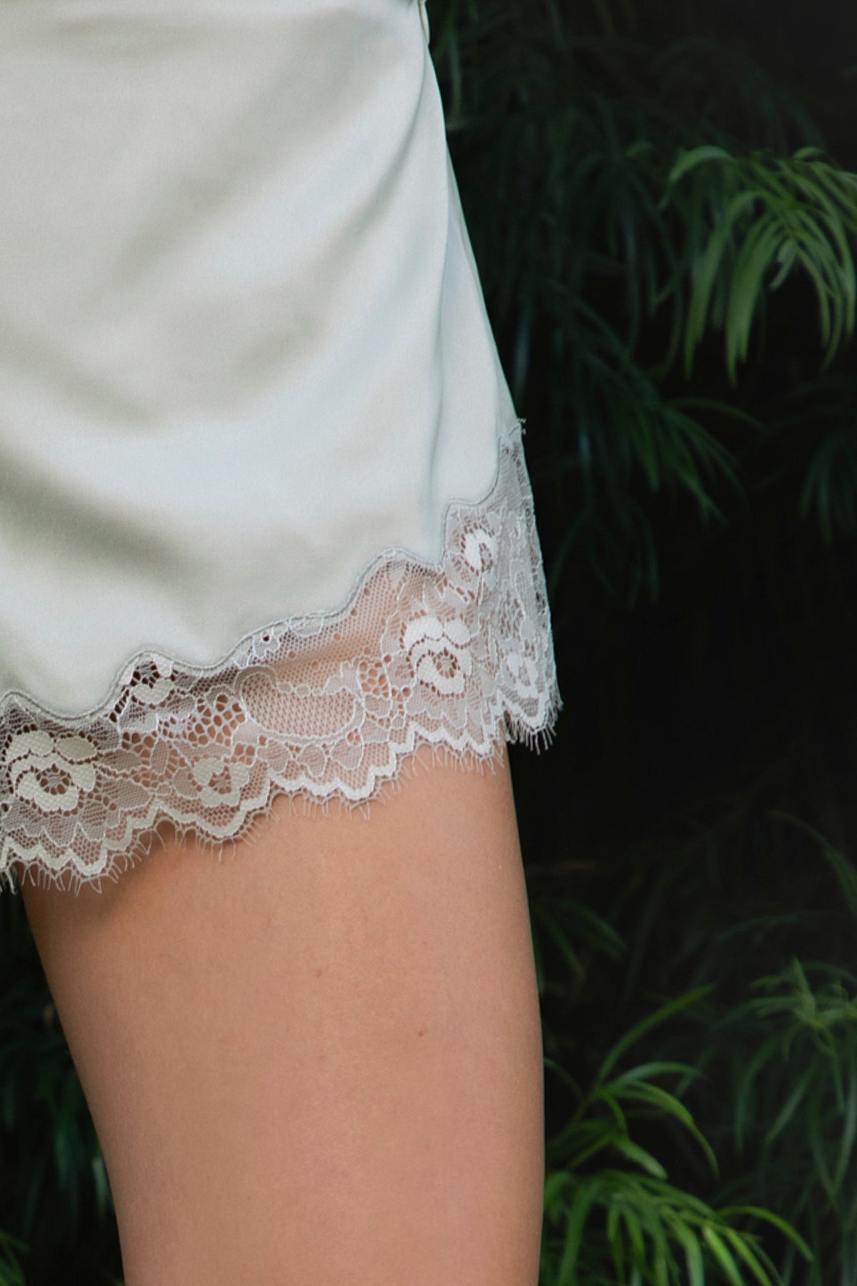 Woman wearing LUNETTE scalloped eyelash lace trim satin shorts IN SAGE featuring soft stretch satin fabric, high rise waist, clean tailored finish, delicate scalloped eyelash lace trim along the hem, hidden back zipper closure, and fully lined interior for added comfort and coverage.