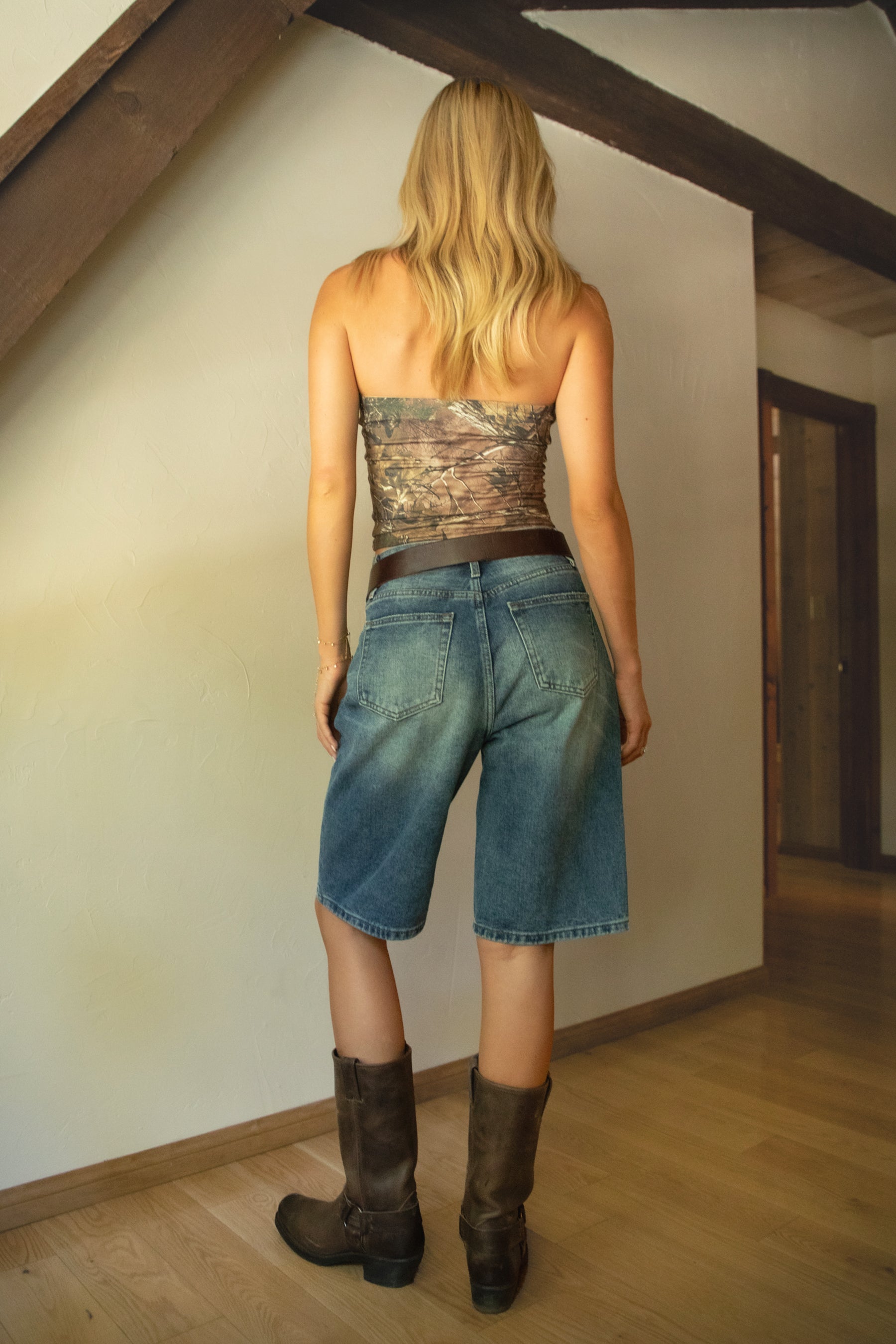 Person wearing a camouflage top, denim shorts, and brown boots in a room with wooden beams.
