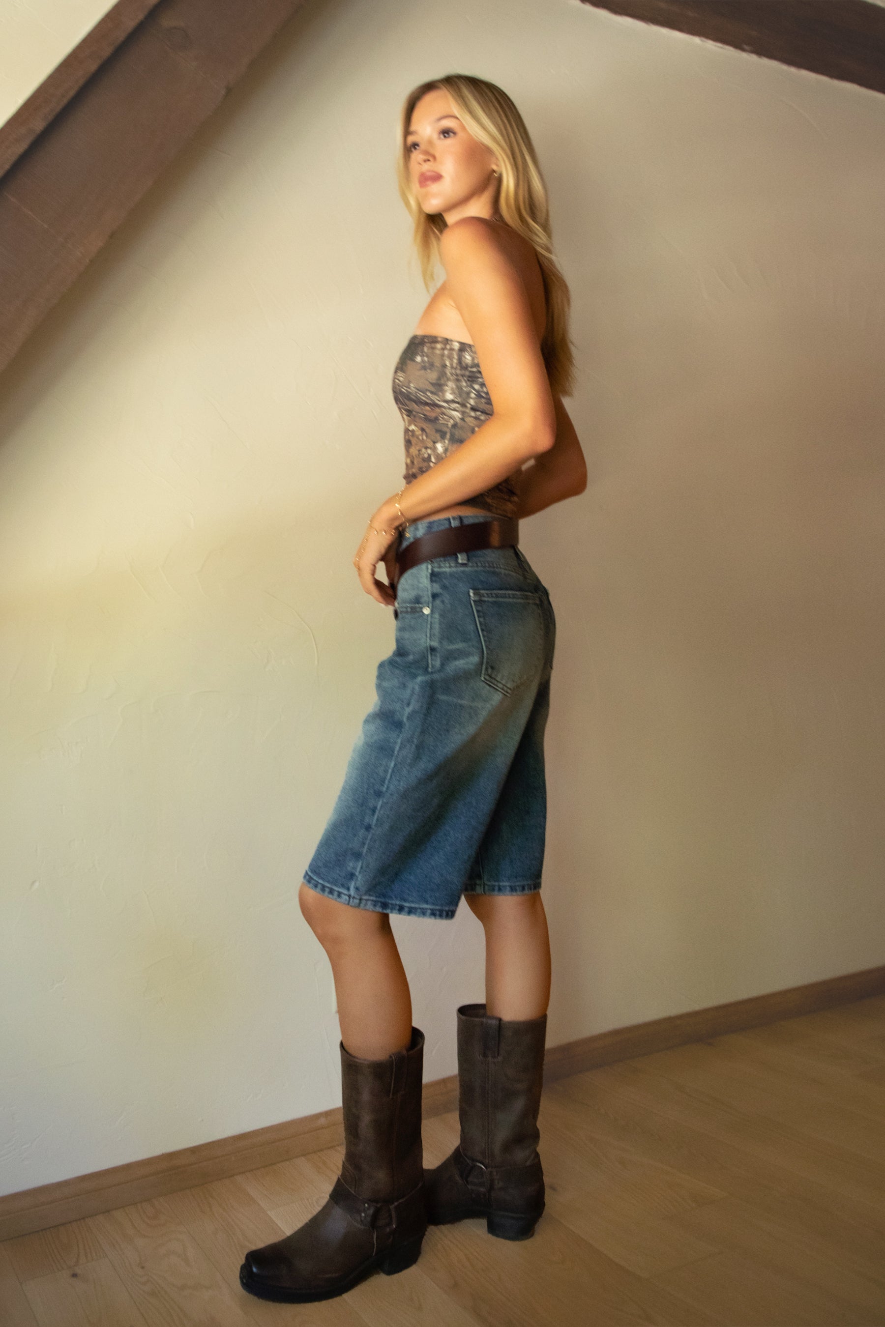 Woman wearing a patterned top, denim skirt, and brown boots standing against a plain wall.