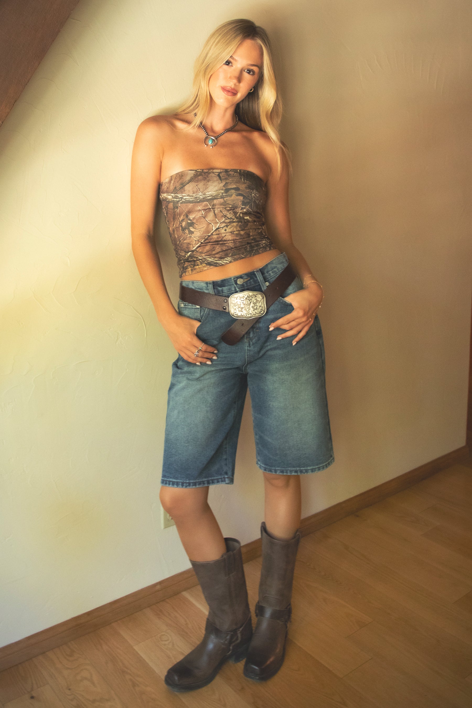 Woman wearing a camouflage top and denim shorts with a belt buckle, standing against a plain wall.