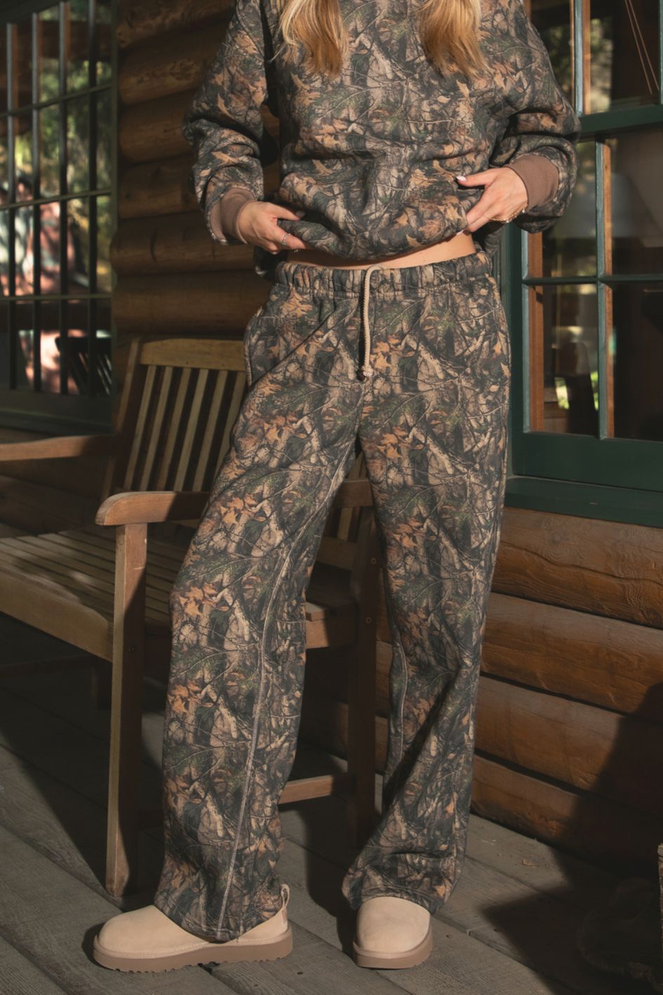 Woman wearing TIC TOC BAYOU Dark Woodland Camo Fleece Wide Leg Sweatpants with deep side pockets, drawcord waist, and drapey Cloud Fleece fabric with matching top on a wooden porch