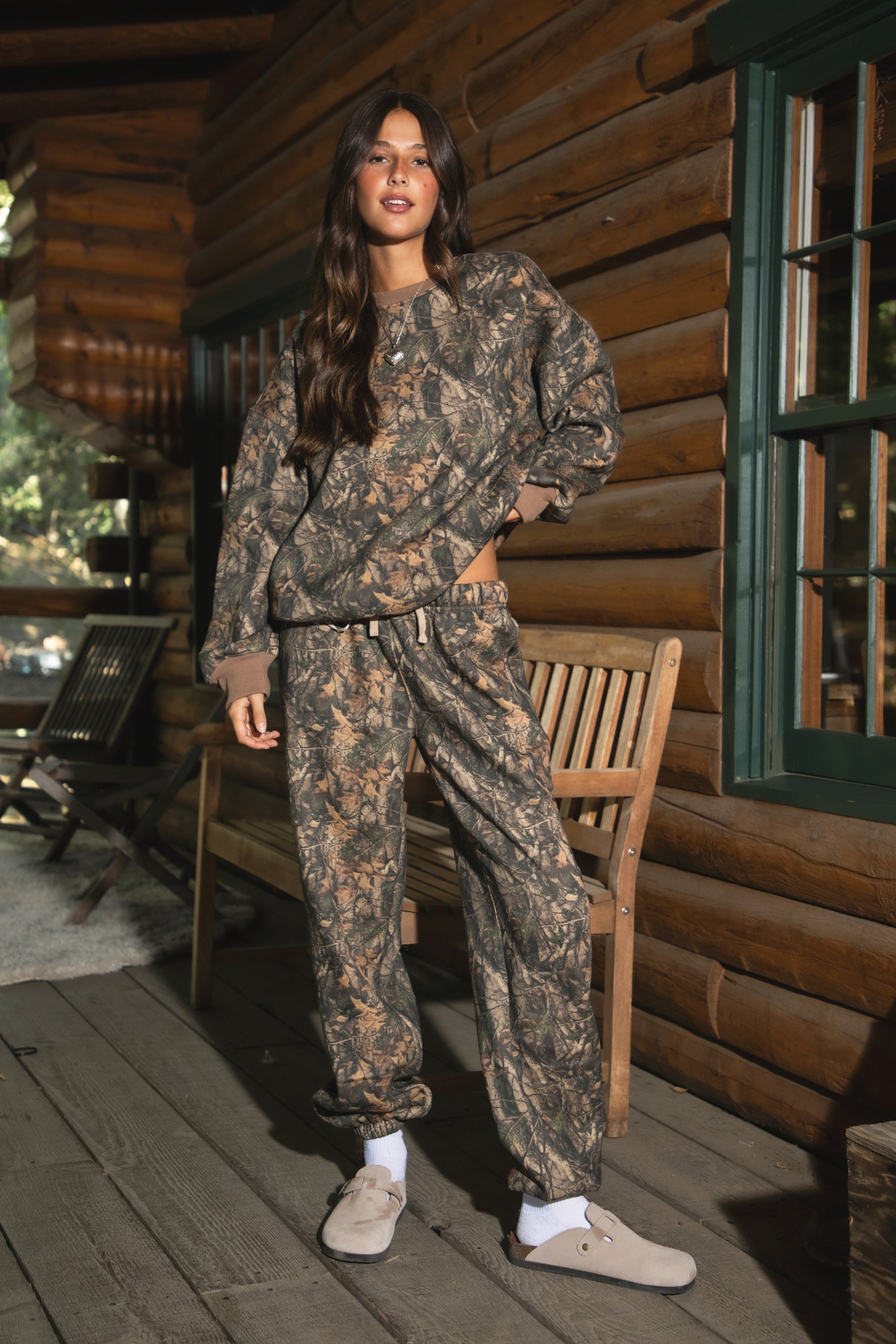 Person wearing TIC TOC BAYOU Dark Woodland Camo Lounge Sweatpants with elastic self-tie waistband, tapered legs, and cinched cuffs in soft Cloud Fleece standing on a wooden deck