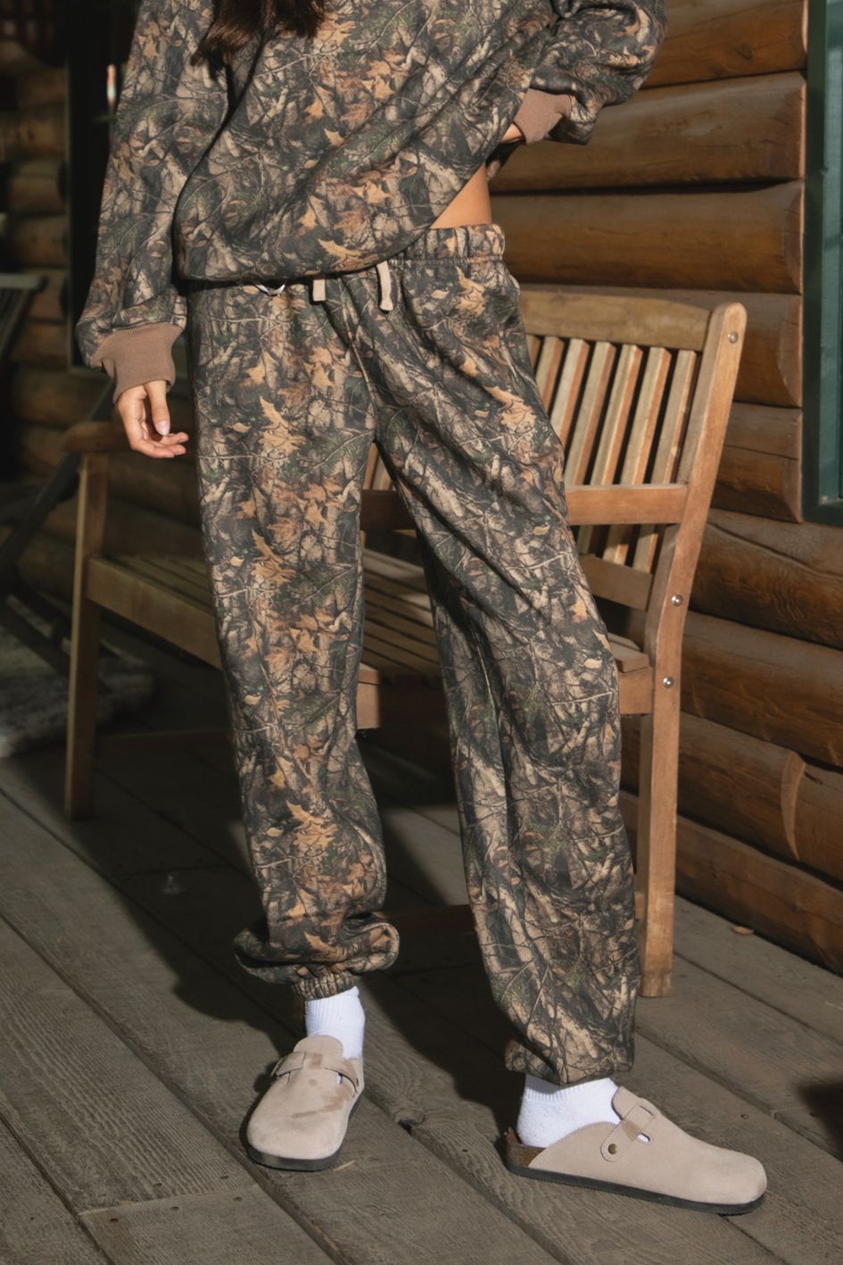 Person wearing TIC TOC BAYOU Dark Woodland Camo Lounge Sweatpants with elastic self-tie waistband, tapered legs, and cinched cuffs in soft Cloud Fleece standing on a wooden deck