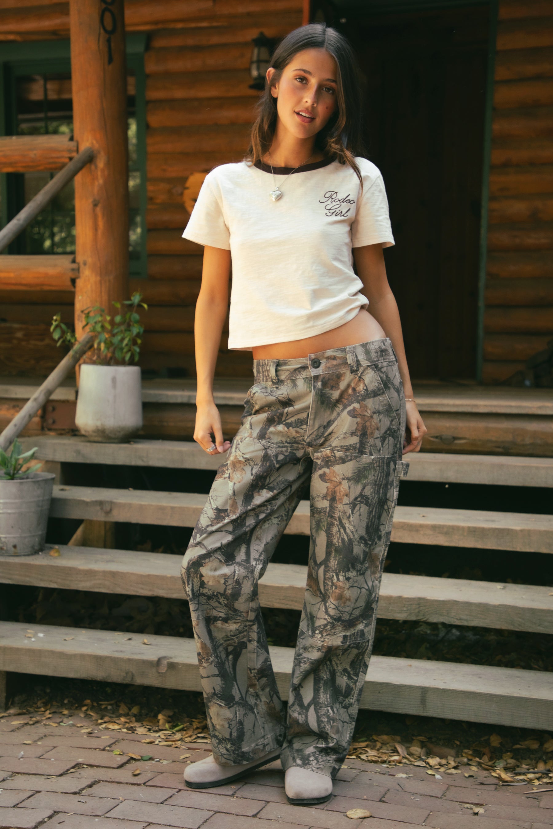 Woman wearing a white t-shirt and TIC TOC WILDWOOD Low Rise Camo Carpenter Pants with wide leg fit, hammer loop, and forest wood camo print.Standing on wooden steps.