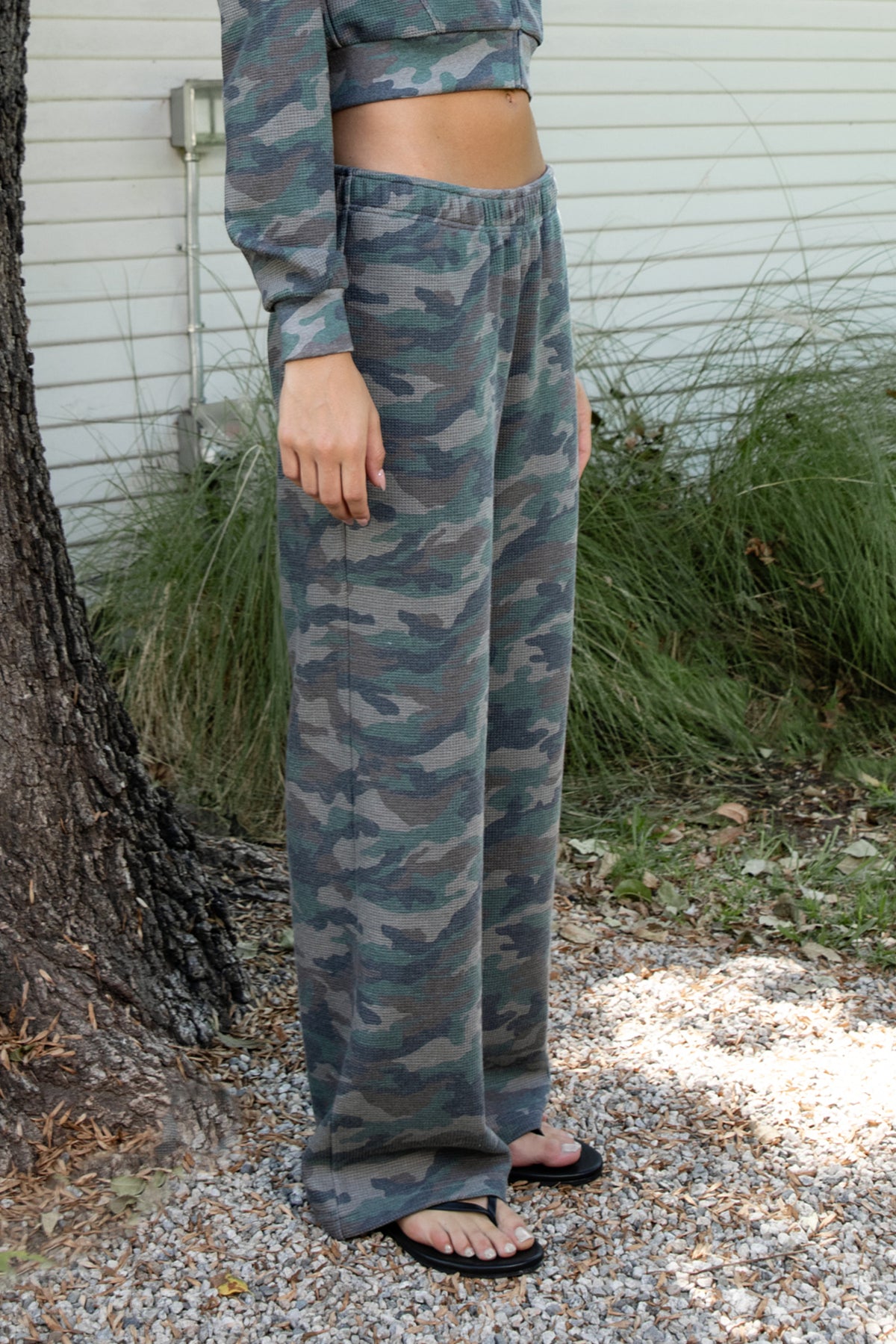 Woman wearing AMAIA heavy weight military camo wide leg pants featuring heavyweight waffle knit fabric with a soft textured feel, allover woodland camouflage print in muted green and grey tones, comfortable elastic waistband, high rise pull on design, functional side pockets, relaxed wide leg silhouette, full length cut, and clean open hem.