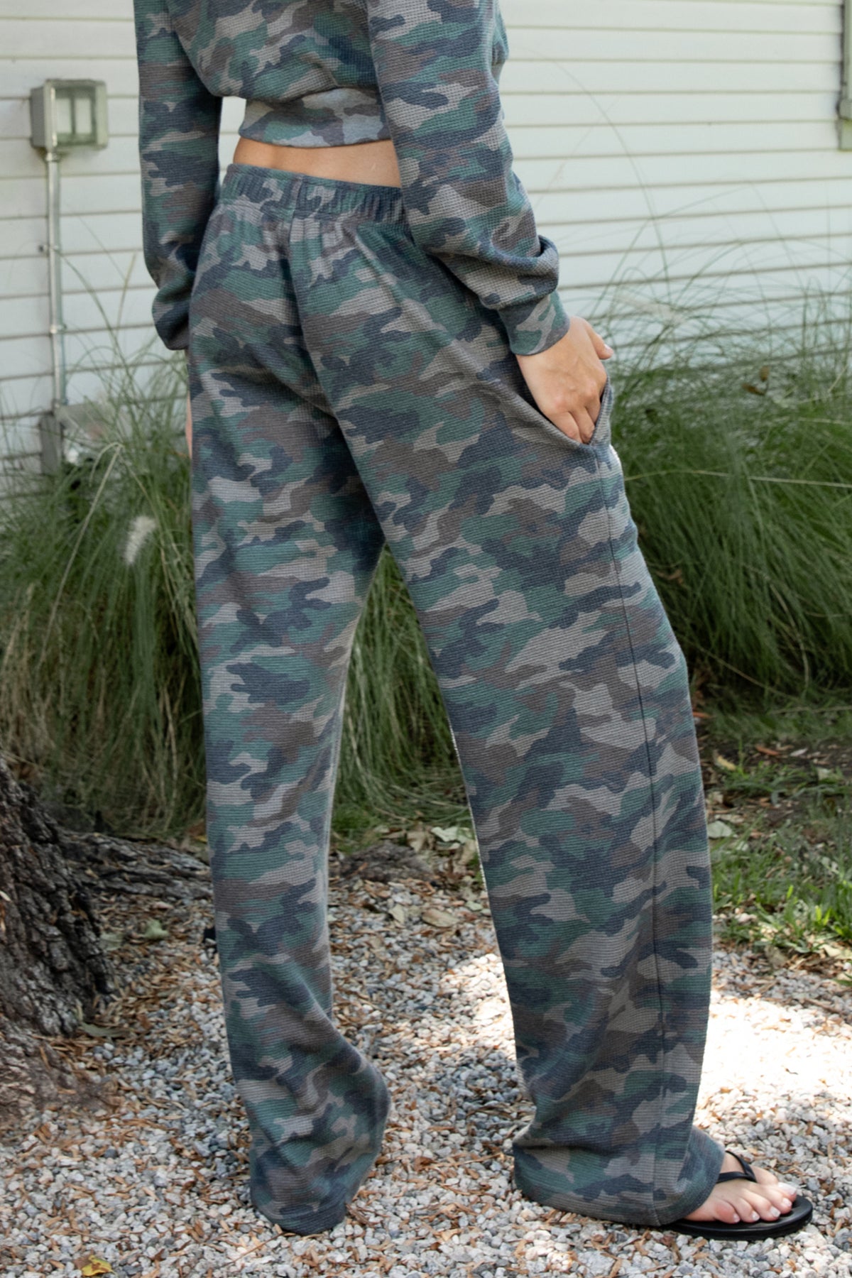 Woman wearing AMAIA heavy weight military camo wide leg pants featuring heavyweight waffle knit fabric with a soft textured feel, allover woodland camouflage print in muted green and grey tones, comfortable elastic waistband, high rise pull on design, functional side pockets, relaxed wide leg silhouette, full length cut, and clean open hem.