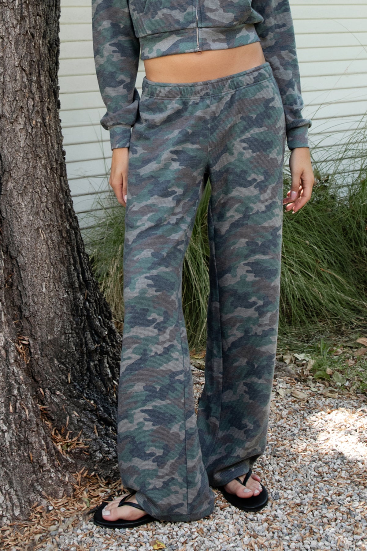 Woman wearing AMAIA heavy weight military camo wide leg pants featuring heavyweight waffle knit fabric with a soft textured feel, allover woodland camouflage print in muted green and grey tones, comfortable elastic waistband, high rise pull on design, functional side pockets, relaxed wide leg silhouette, full length cut, and clean open hem.
