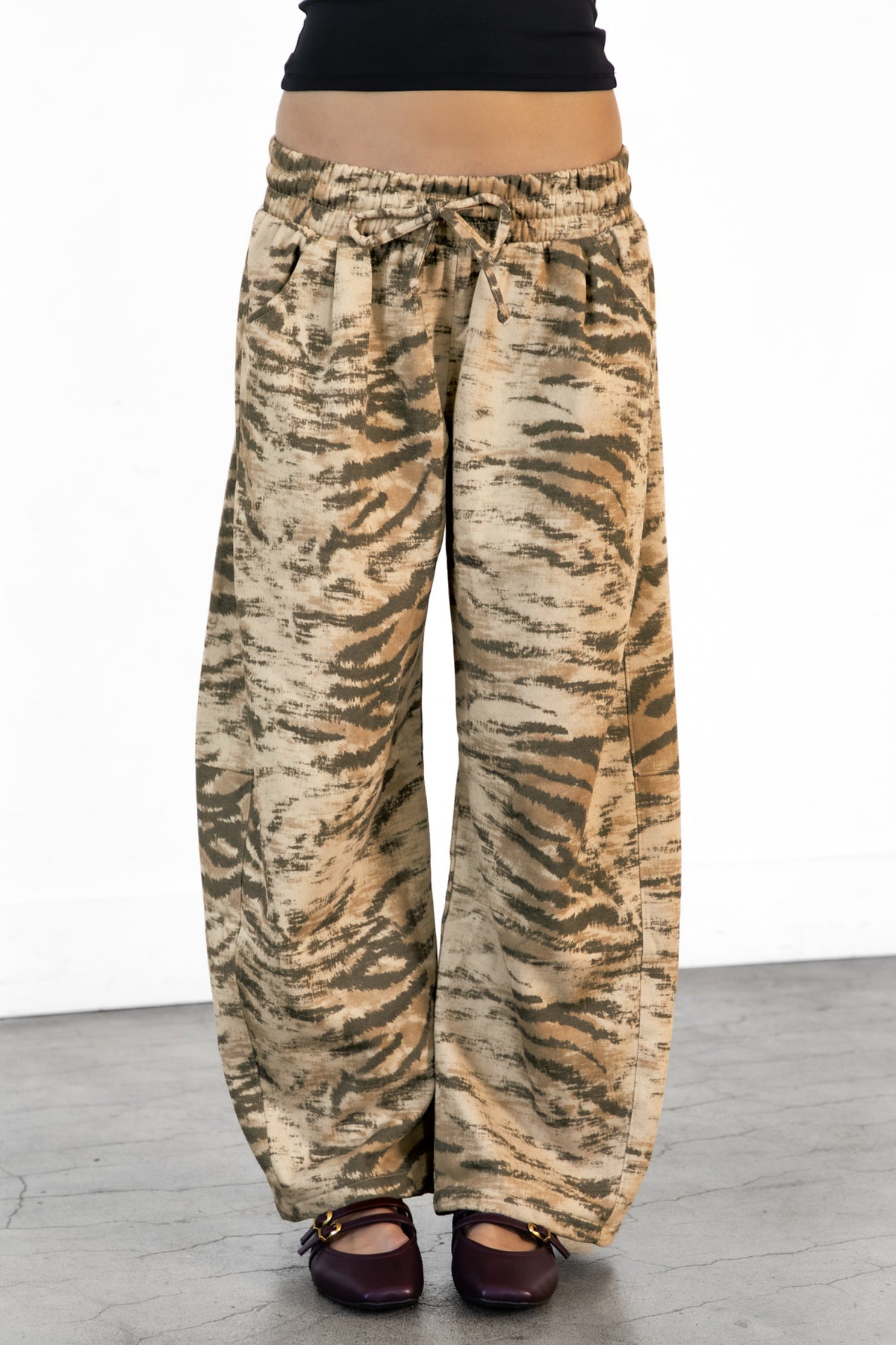 Woman wearing ZARIA tiger print wide leg pants featuring soft polyester cotton blend fabric, allover abstract tiger print in earthy beige, brown, and olive tones, elastic waistband with adjustable drawstring tie, relaxed fit through the hips and thighs, functional side pockets, wide leg silhouette with a subtle barrel shape, and full length design with paneled seam detailing.