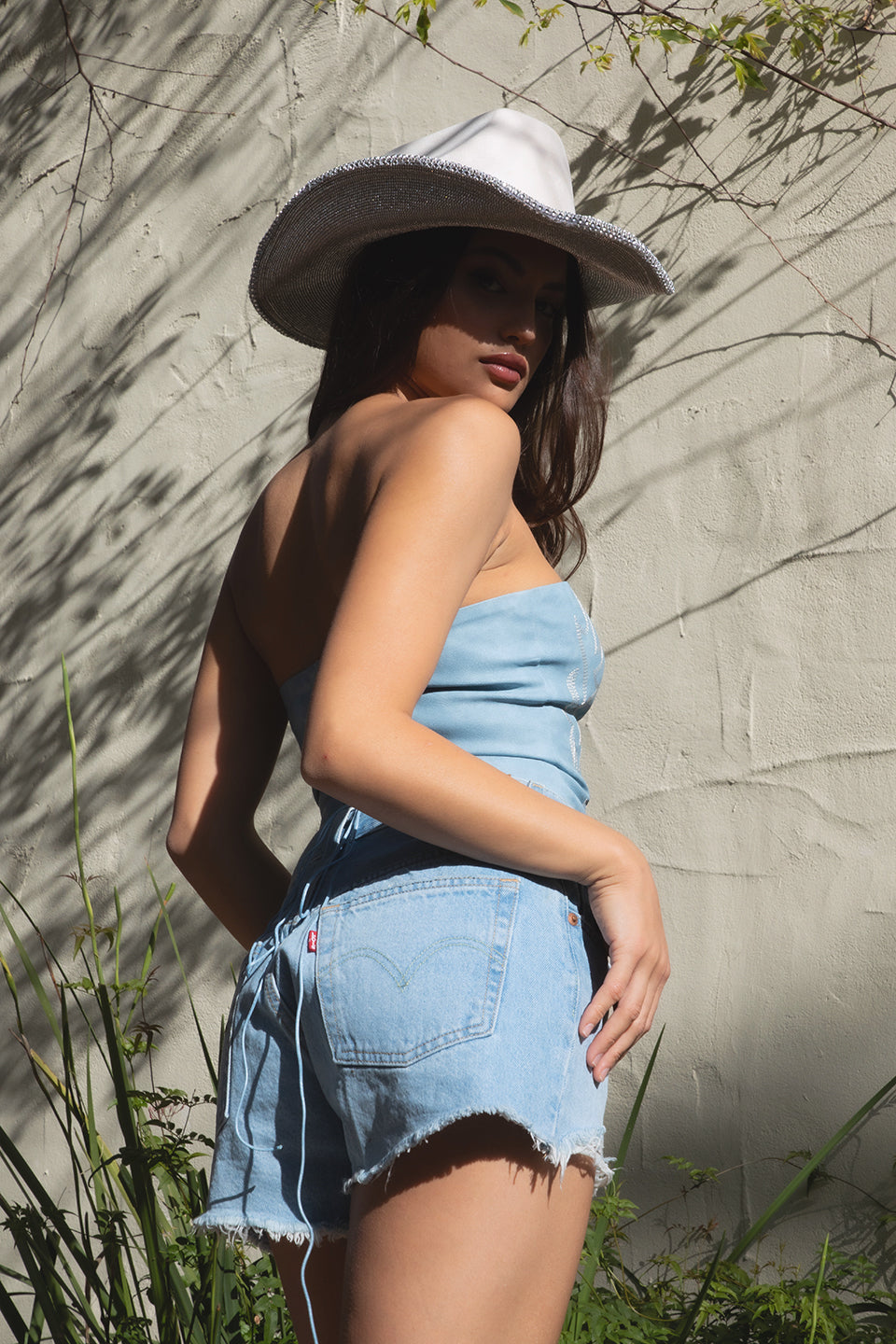 Woman wearing a denim blue strapless romper and wide-brimmed hat against a stone wall.