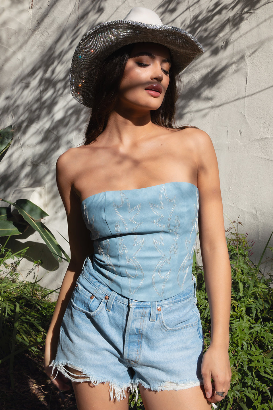 Woman wearing a denim blue strapless romper and wide-brimmed hat against a stone wall.