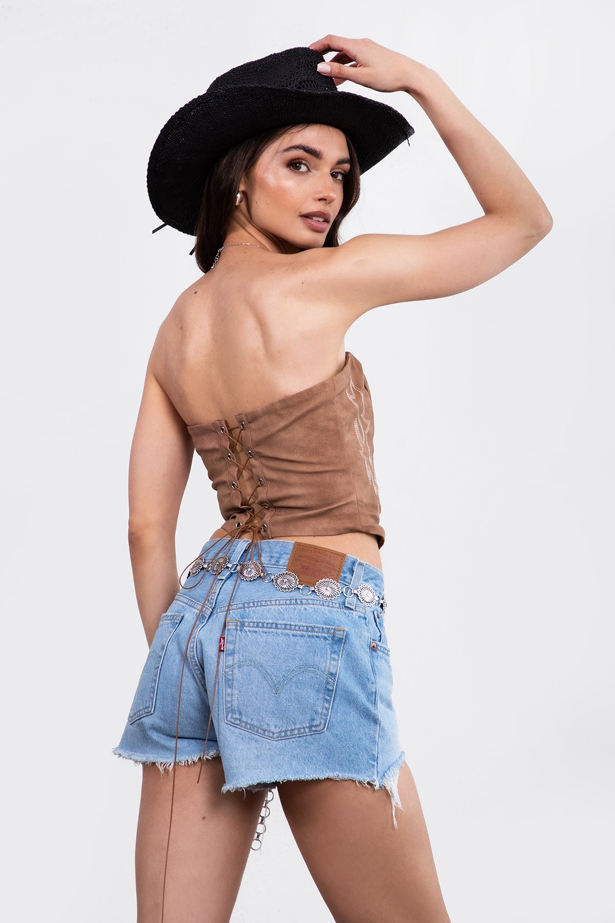 A woman wearing a strapless, boned panel construction light brown top with embroidered details and a straight neckline. matching with denim shorts and wide rim black cow boy hat