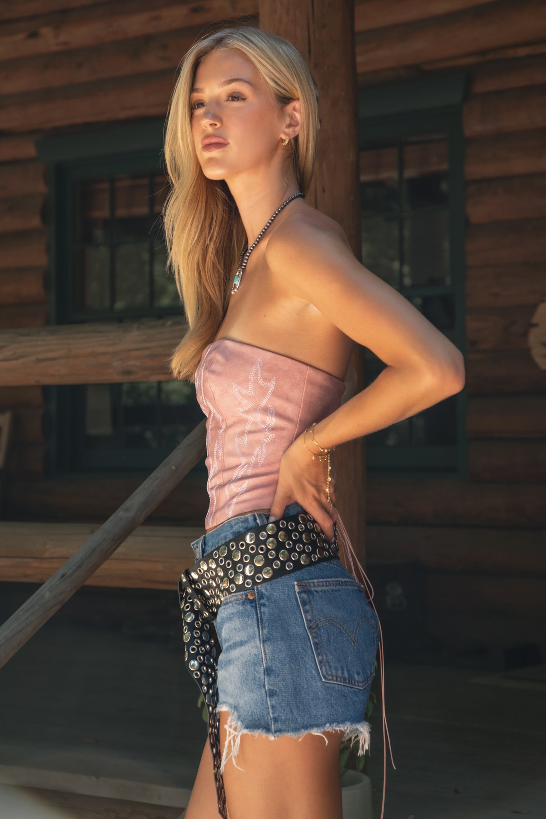 Woman wearing a pink embroidered western top and denim shorts with a studded belt, standing in front of a wooden wall.
