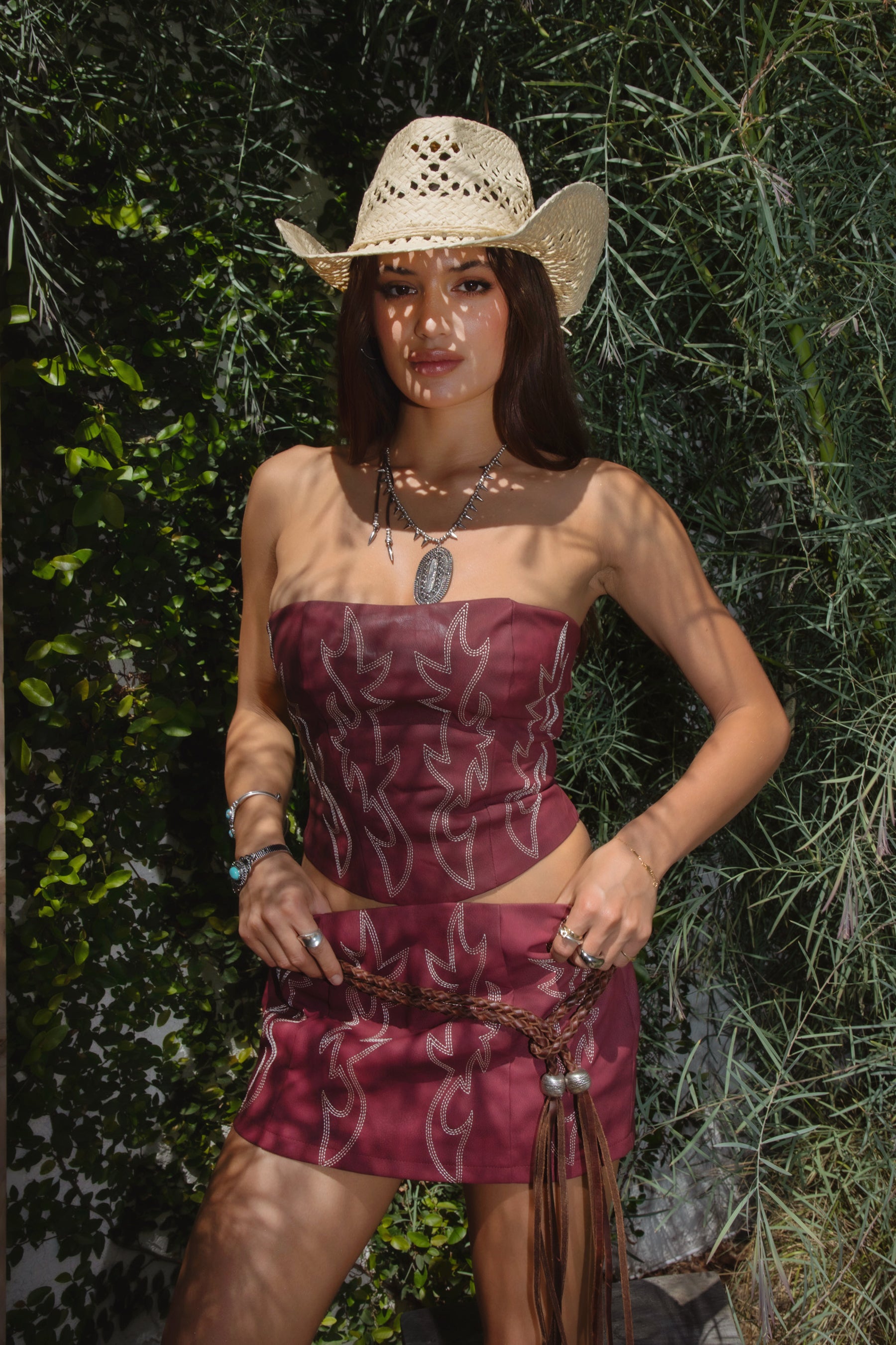 A woman wearing a red strapless, boned panel construction top with embroidered western details and a straight neckline with matching mini skirt, straw hat, and western accessories. 