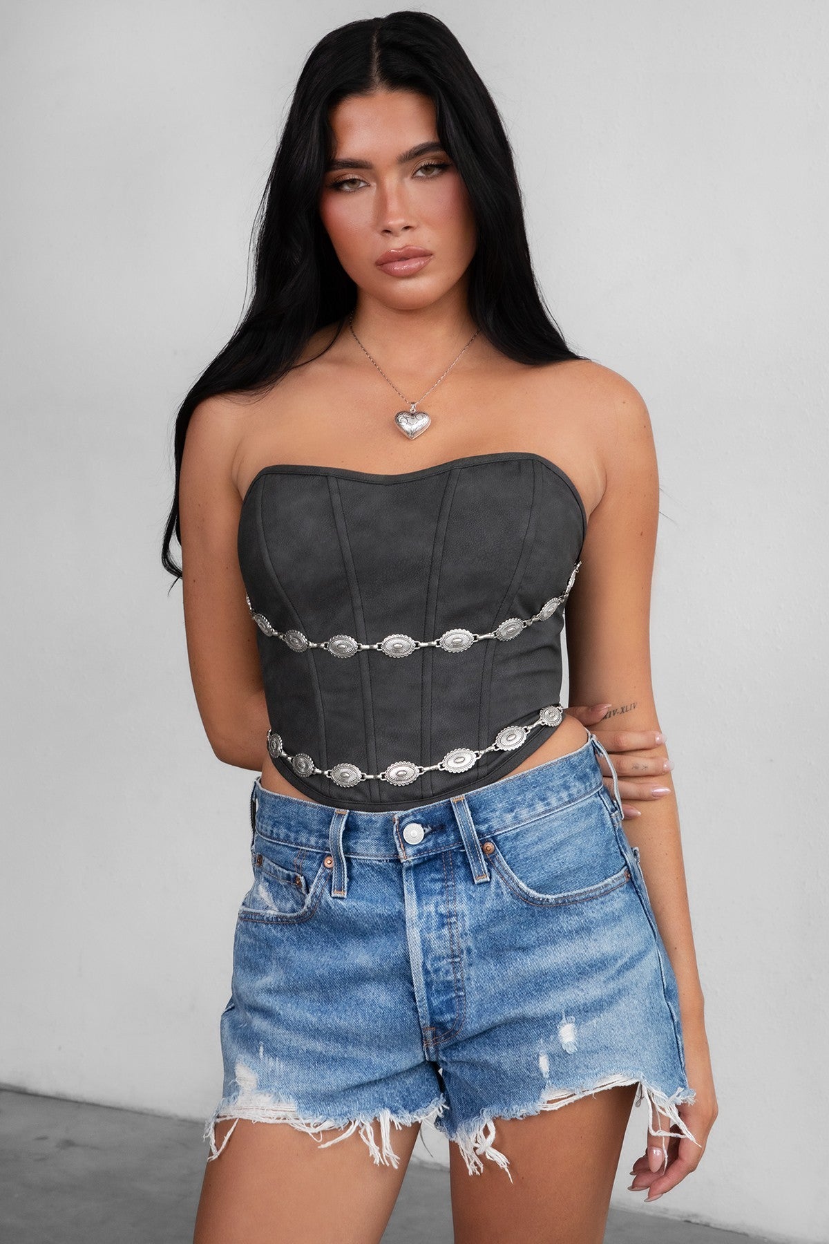 Woman wearing TIC TOC GLENDORA Distressed PU Leather Corset Top in black with strapless sweetheart neckline and silver western embellishments paired with blue denim shorts.