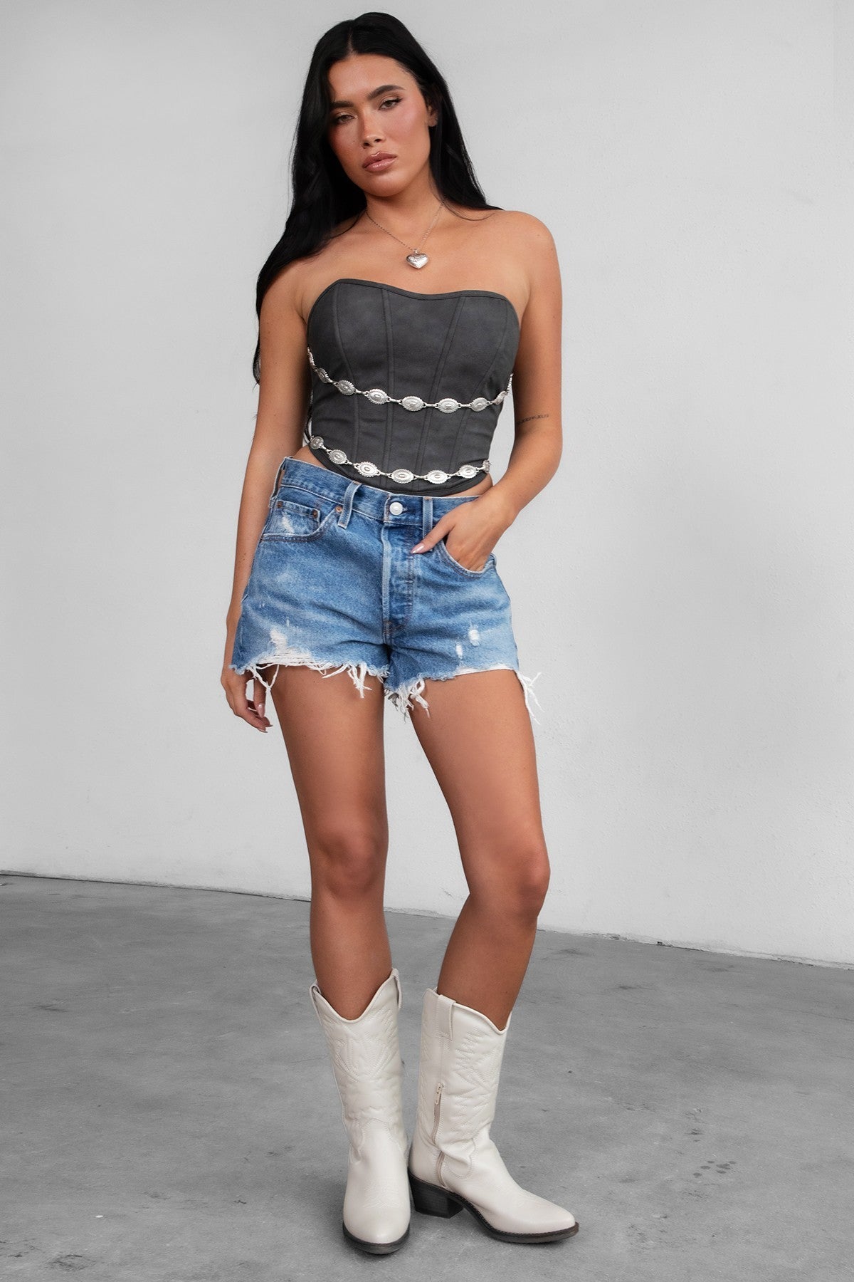 Woman wearing TIC TOC GLENDORA Distressed PU Leather Corset Top in black with strapless sweetheart neckline and silver western embellishments paired with blue denim shorts.