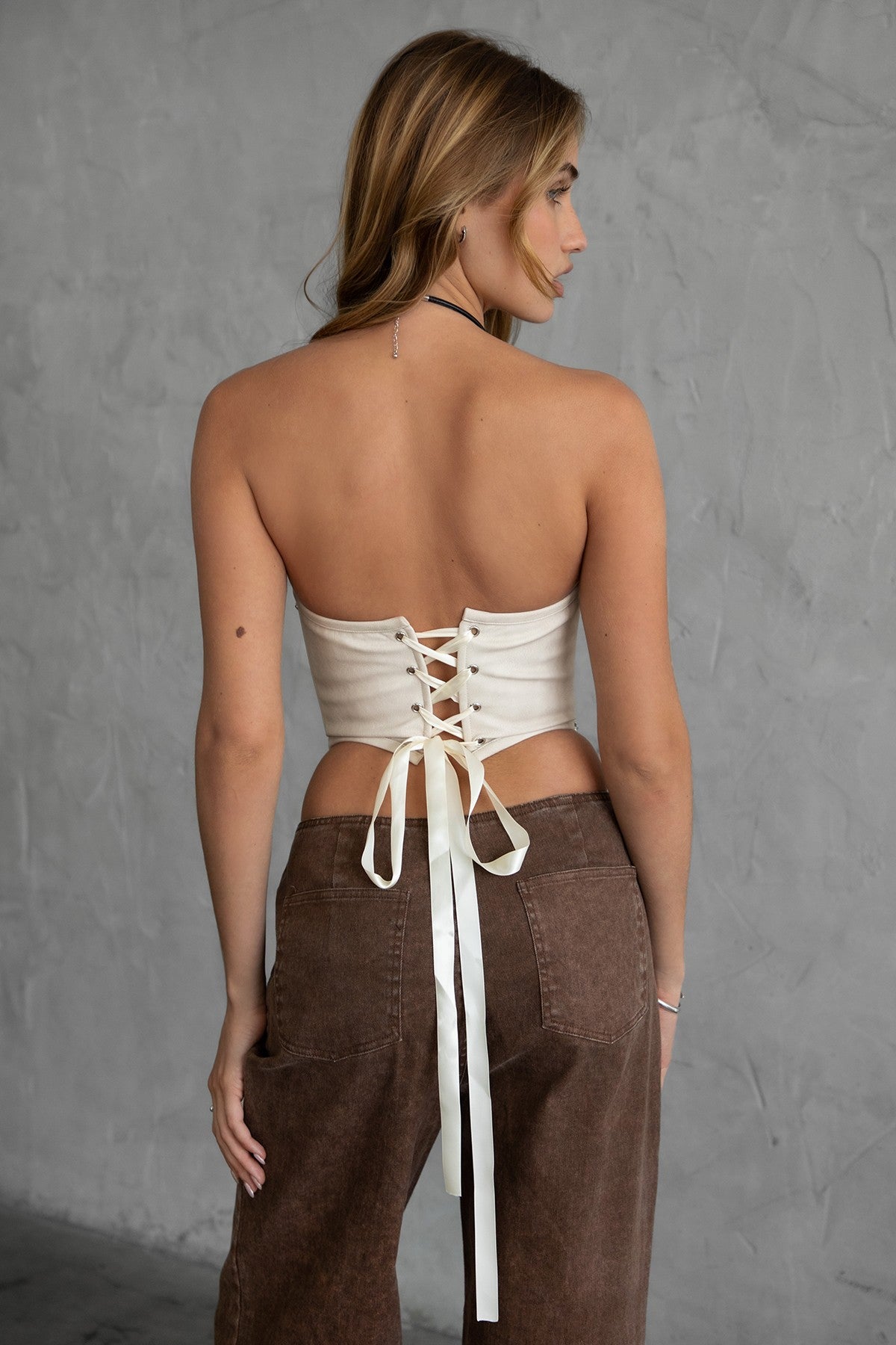 Woman wearing TIC TOC GLENDORA Distressed PU Leather Corset Top in cream with strapless sweetheart neckline and silver western embellishments paired with brown pants.