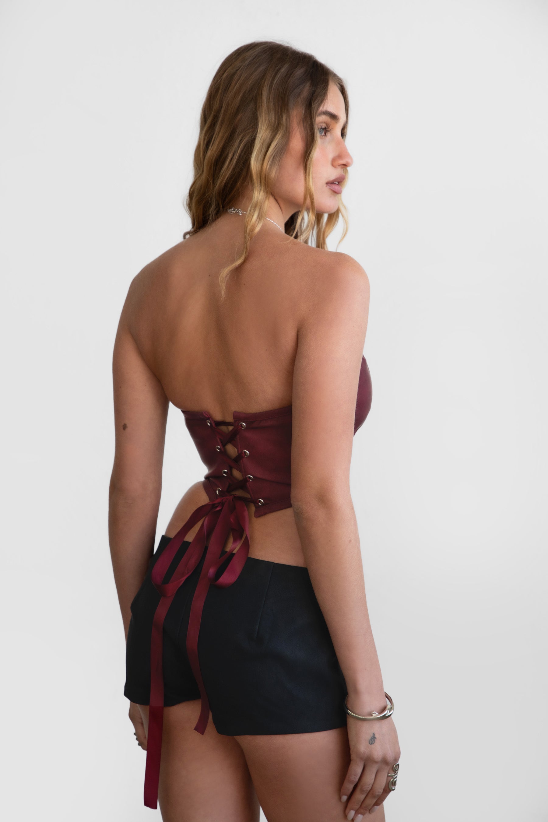 Woman wearing TIC TOC GLENDORA Distressed PU Leather Corset Top in red with strapless sweetheart neckline and silver western embellishments paired with blacl shorts.