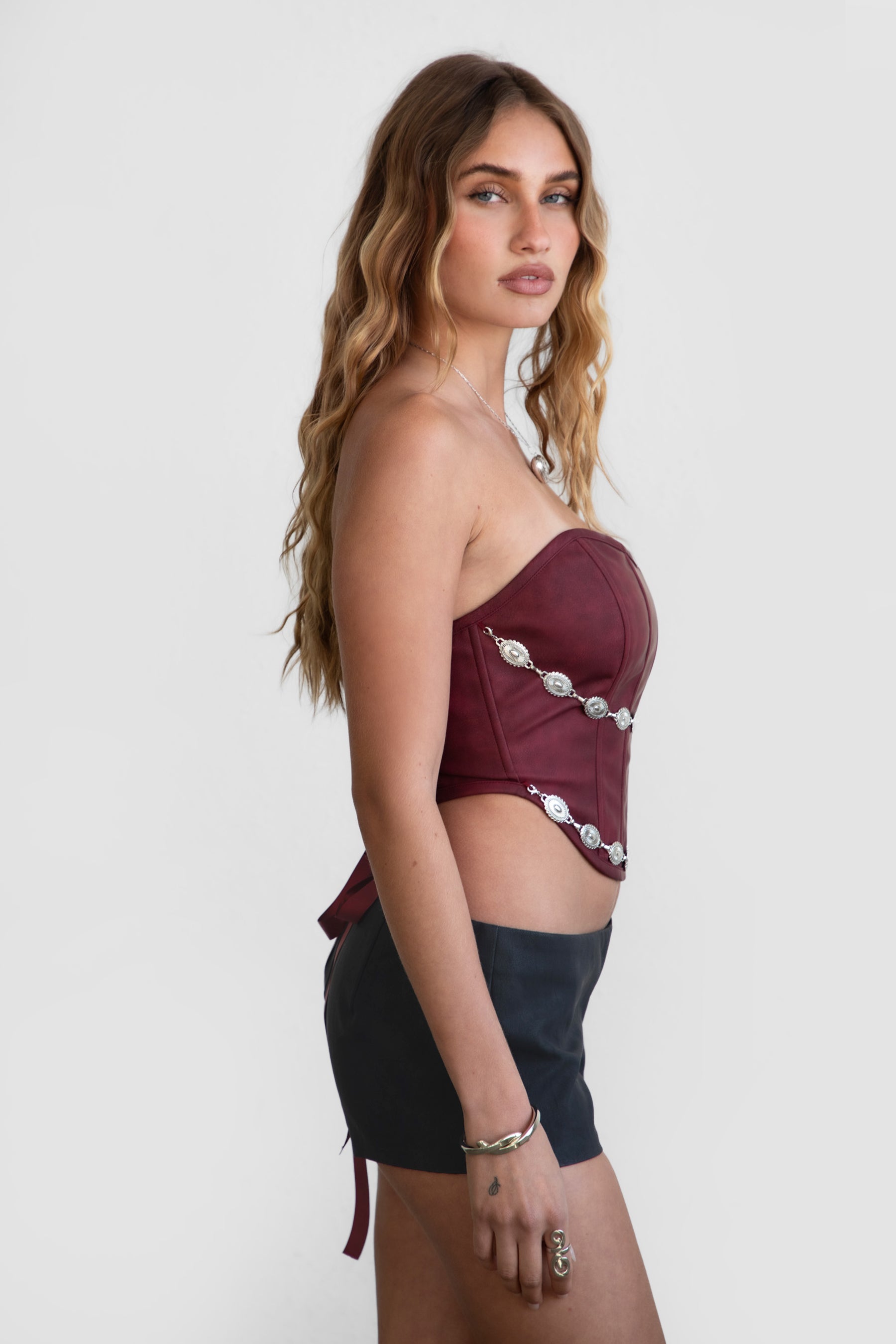Woman wearing TIC TOC GLENDORA Distressed PU Leather Corset Top in red with strapless sweetheart neckline and silver western embellishments paired with blacl shorts.