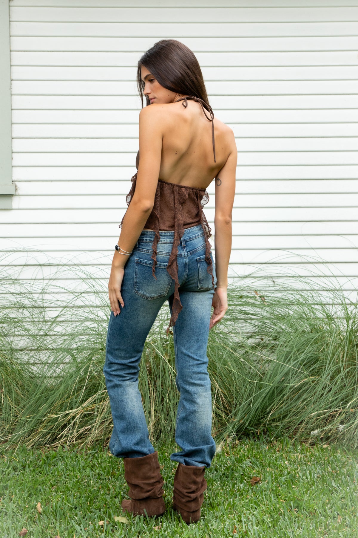 Woman wearing ZARELA faux leather lace ruffle halter top featuring a deep V neckline with metal eyelet detailing, adjustable halter neck tie closure, faux leather triangle bust design, low cut open back silhouette, sheer floral lace contrast body, asymmetrical cascading lace ruffle detailing, and cropped length with a flowing lace hem.