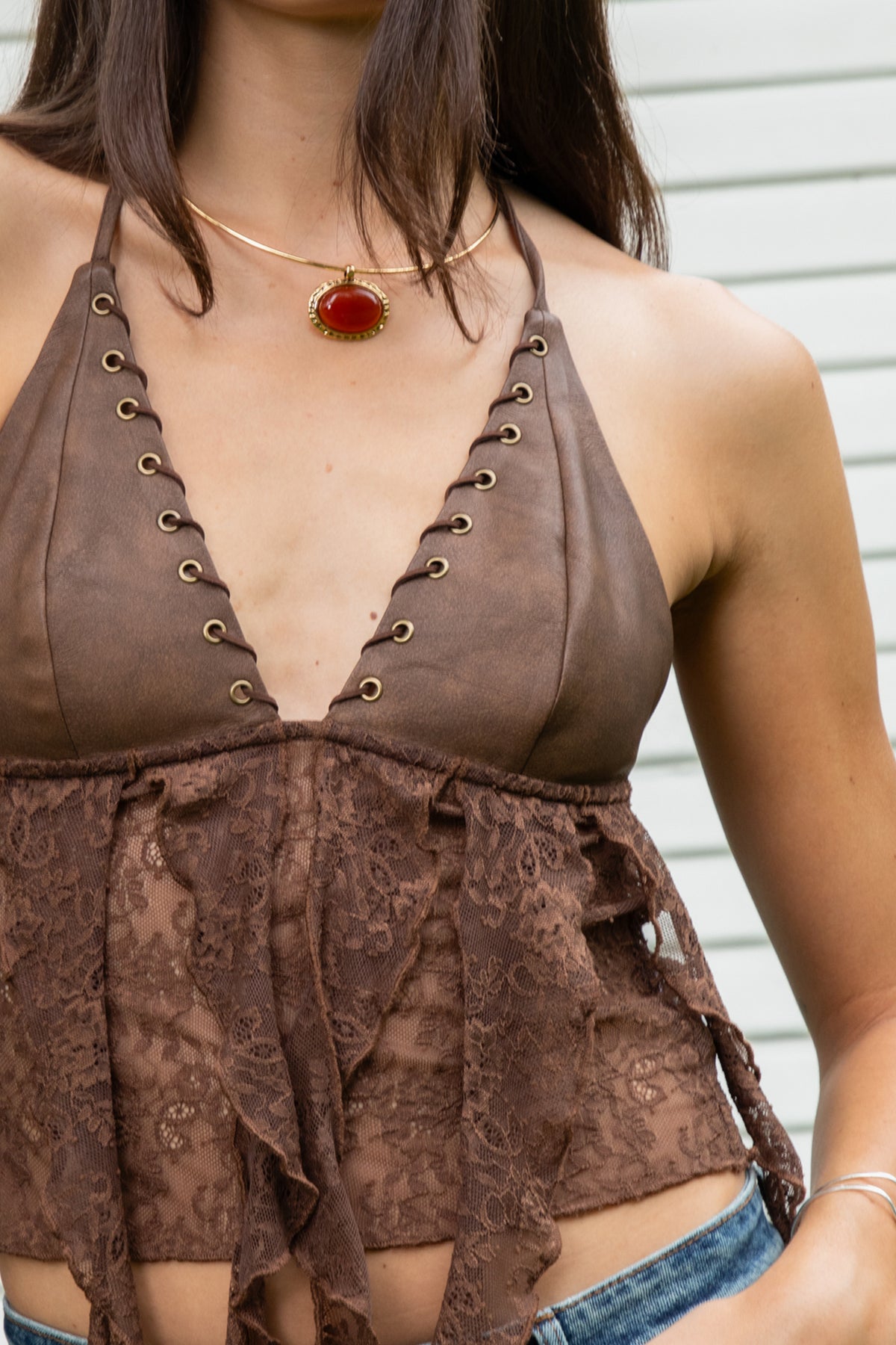 Woman wearing ZARELA faux leather lace ruffle halter top featuring a deep V neckline with metal eyelet detailing, adjustable halter neck tie closure, faux leather triangle bust design, low cut open back silhouette, sheer floral lace contrast body, asymmetrical cascading lace ruffle detailing, and cropped length with a flowing lace hem.