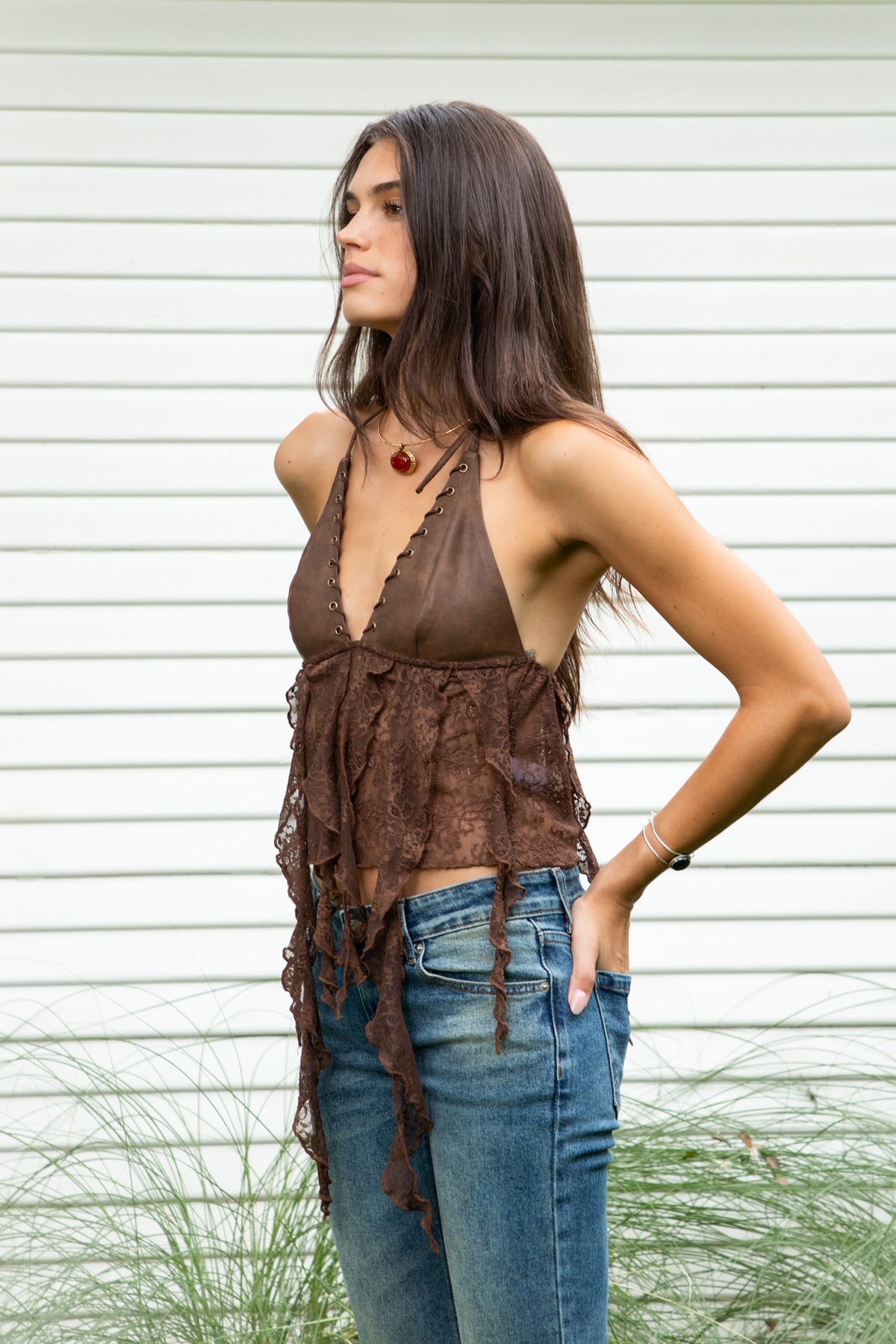 Woman wearing ZARELA faux leather lace ruffle halter top featuring a deep V neckline with metal eyelet detailing, adjustable halter neck tie closure, faux leather triangle bust design, low cut open back silhouette, sheer floral lace contrast body, asymmetrical cascading lace ruffle detailing, and cropped length with a flowing lace hem.