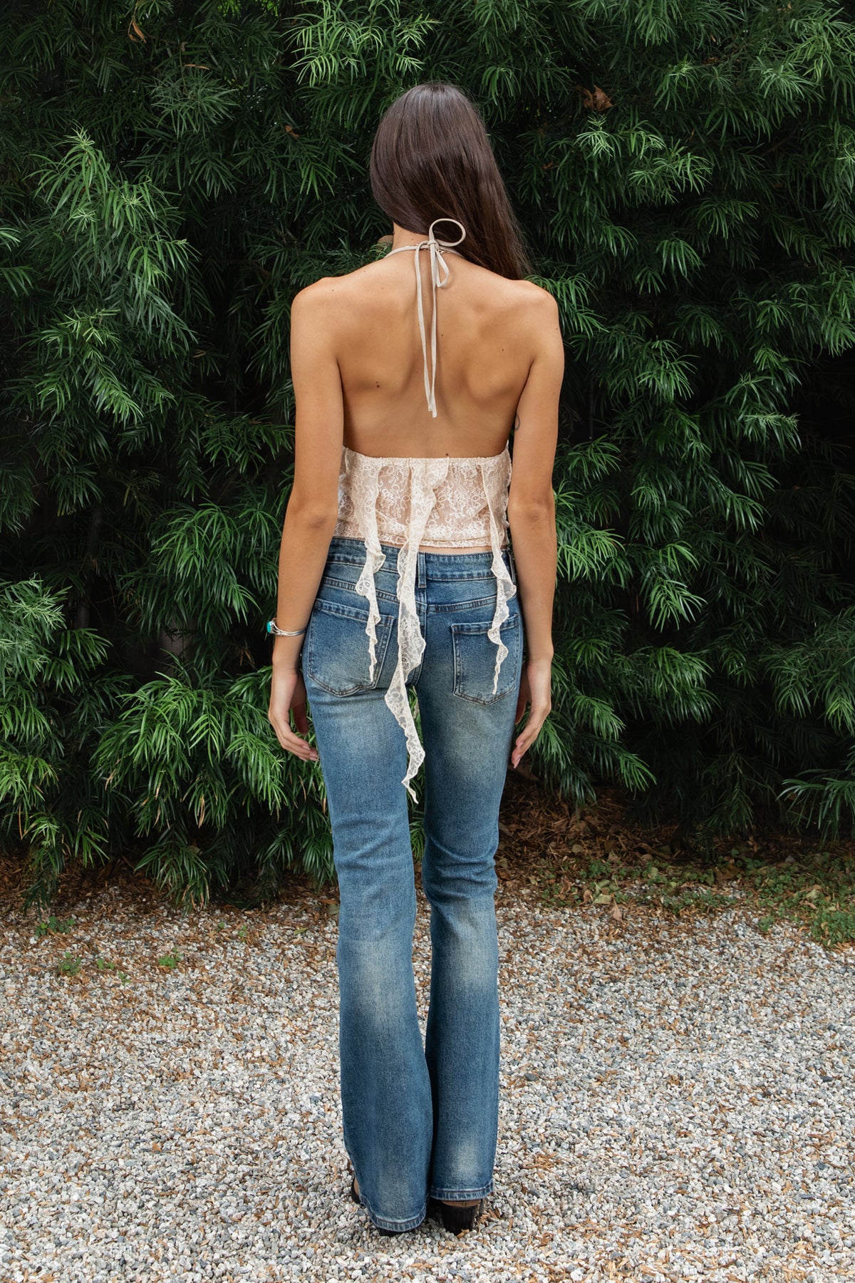Woman wearing ZARELA faux leather lace ruffle halter top featuring a deep V neckline with metal eyelet detailing, adjustable halter neck tie closure, faux leather triangle bust design, low cut open back silhouette, sheer floral lace contrast body, asymmetrical cascading lace ruffle detailing, and cropped length with a flowing lace hem.
