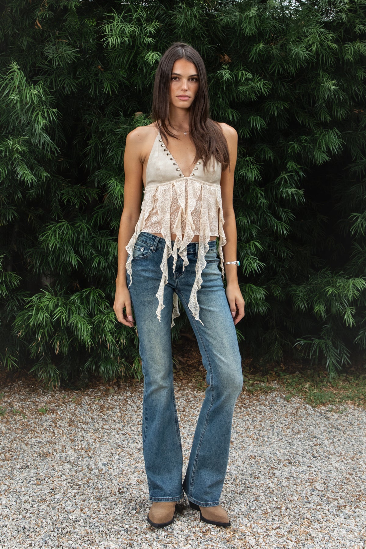 Woman wearing ZARELA faux leather lace ruffle halter top featuring a deep V neckline with metal eyelet detailing, adjustable halter neck tie closure, faux leather triangle bust design, low cut open back silhouette, sheer floral lace contrast body, asymmetrical cascading lace ruffle detailing, and cropped length with a flowing lace hem.