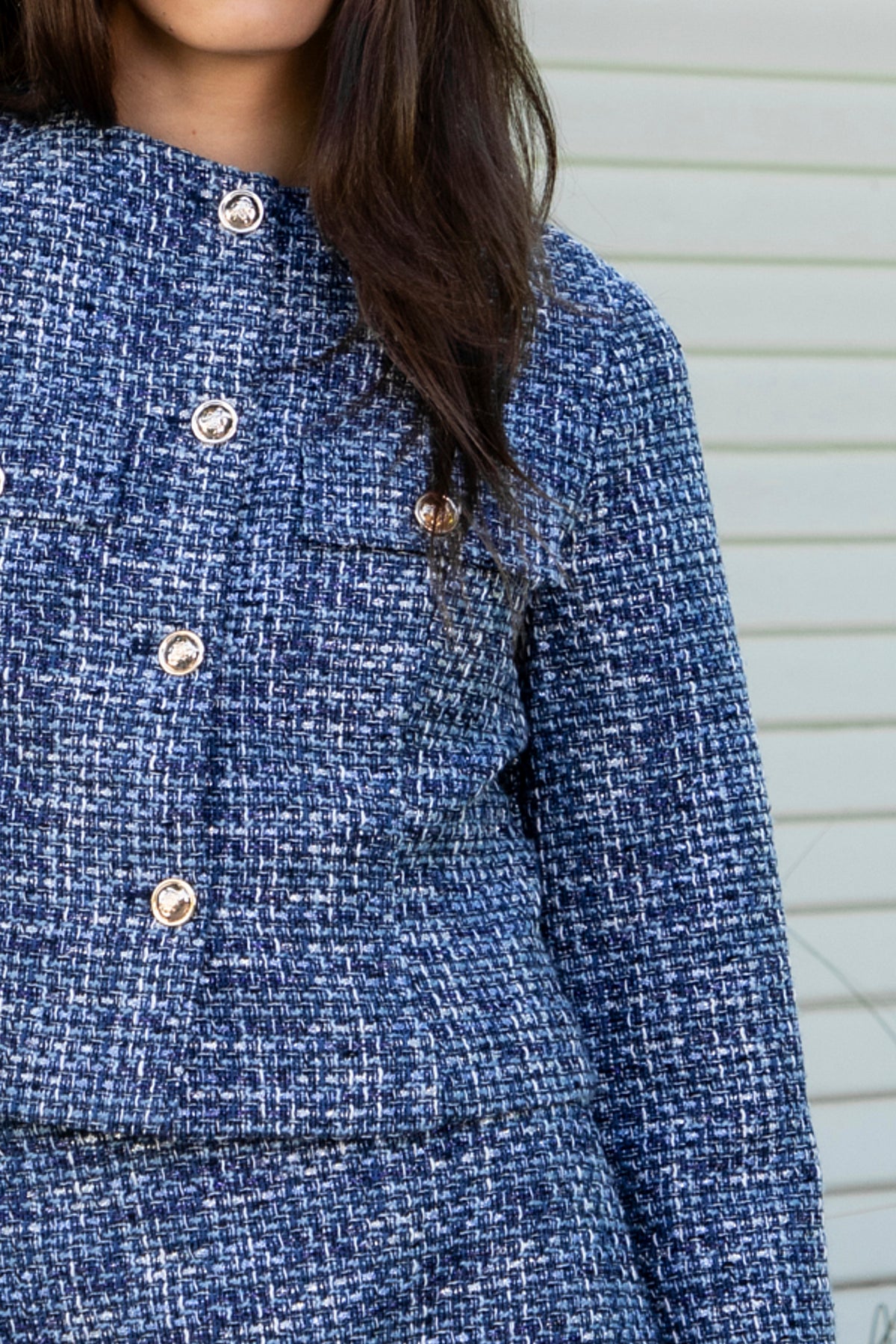 Woman wearing COCO navy blue lurex tweed long sleeve jacket featuring textured lurex tweed fabric, structured cropped silhouette, round neckline, clean collarless design, gold tone statement button front closure, decorative chest flap pockets with matching gold tone buttons, subtle metallic threading, and full lining for a smooth finish.
