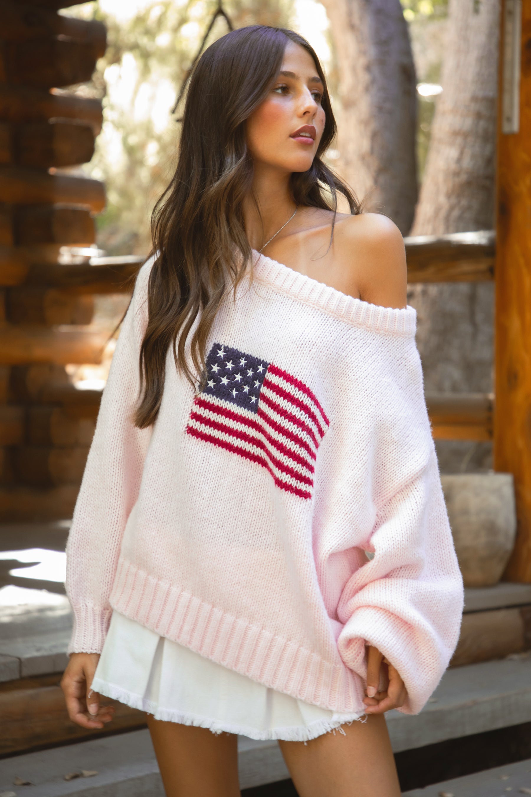 Woman wearing a TIC TOC BRANDI Ribbed Knit Sweater in pink with off-the-shoulder boat neck, American flag pattern, and semi-oversized fit, paired with white denim skirt, standing on wooden steps. 