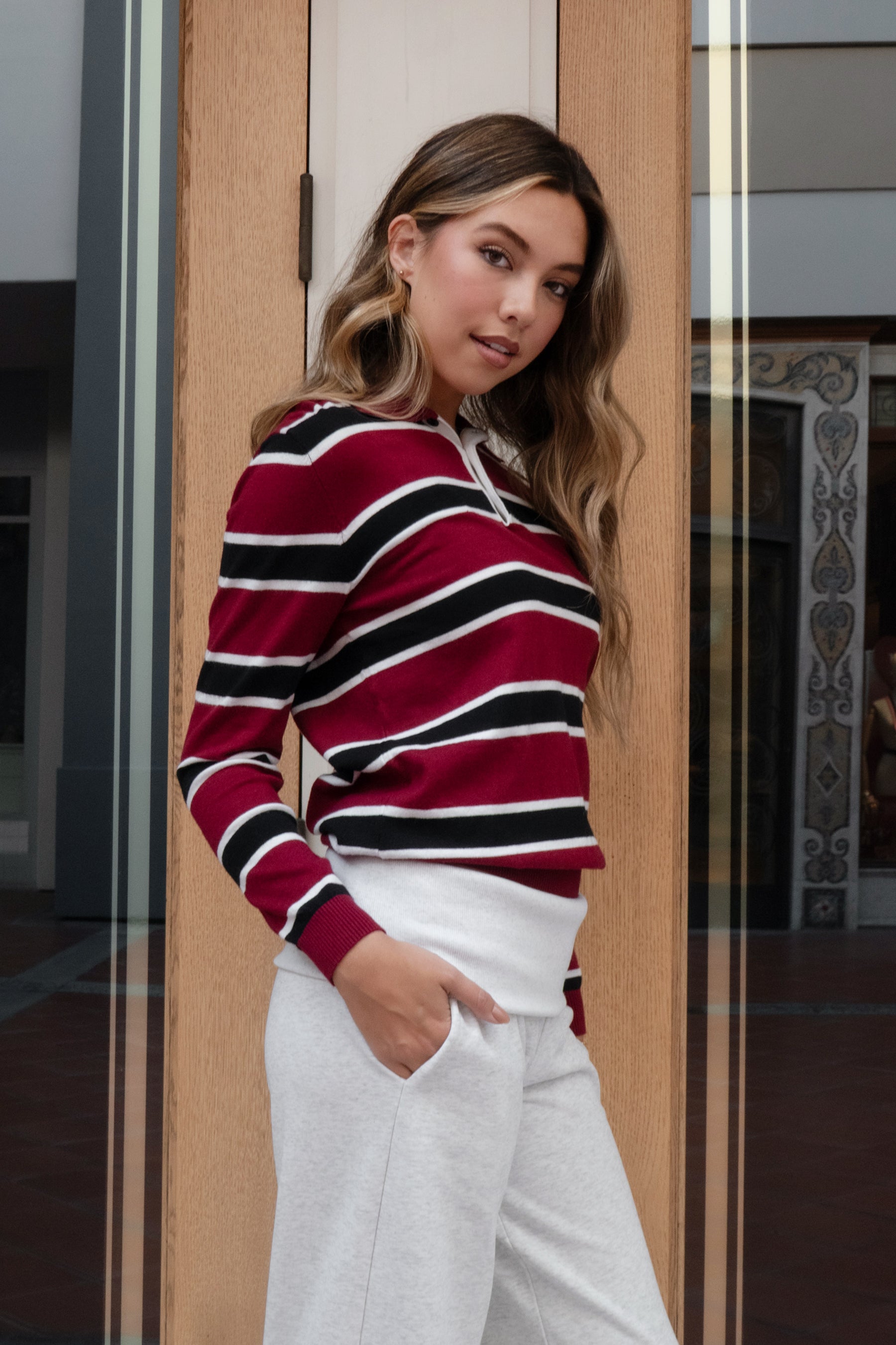 Woman wearing a TIC TOC RHODES Striped Polo T-Shirt red and black striped sweater with white pants, standing indoors.