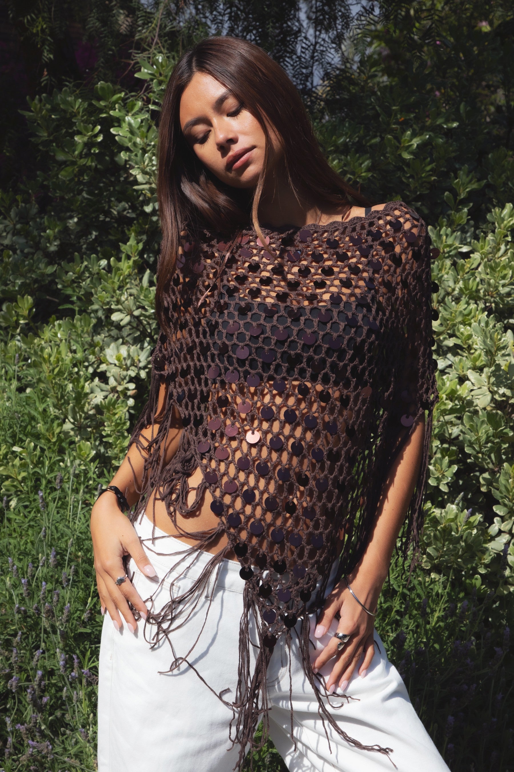 Woman wearing TIC TOC TC31690-LUNARA LUNARA sequin crochet fringe poncho featuring open crochet knit fabric, large allover sequin embellishments, a scalloped wide neckline worn off one shoulder, relaxed poncho silhouette, triangle point hem, and long fringe tassel trim, styled over a fitted top for a western festival and vacation-ready boho look.
