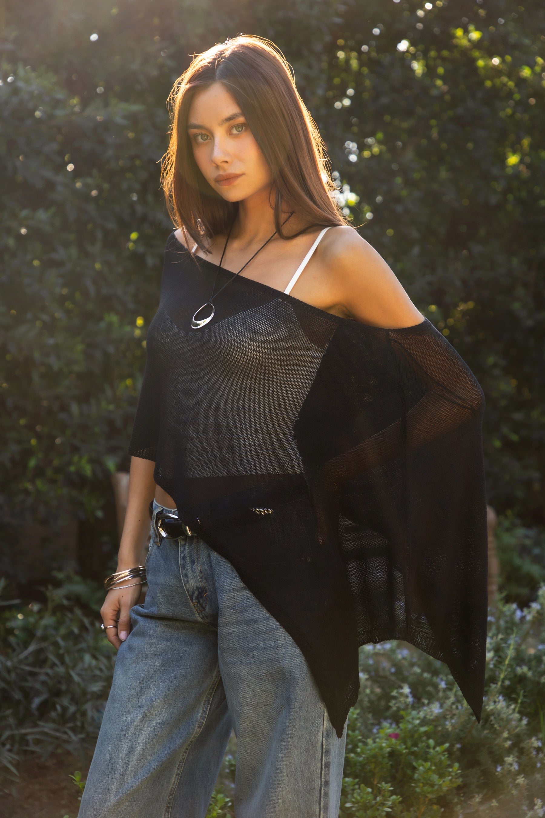 Woman wearing TIC TOC TC31873 SOLEI lightweight open sheer sweater knit poncho in BLACK, featuring a wide off-shoulder neckline, airy open knit texture, relaxed draped poncho silhouette, and asymmetrical hem, styled over a white cami with light wash denim for a western-casual summer layering look.