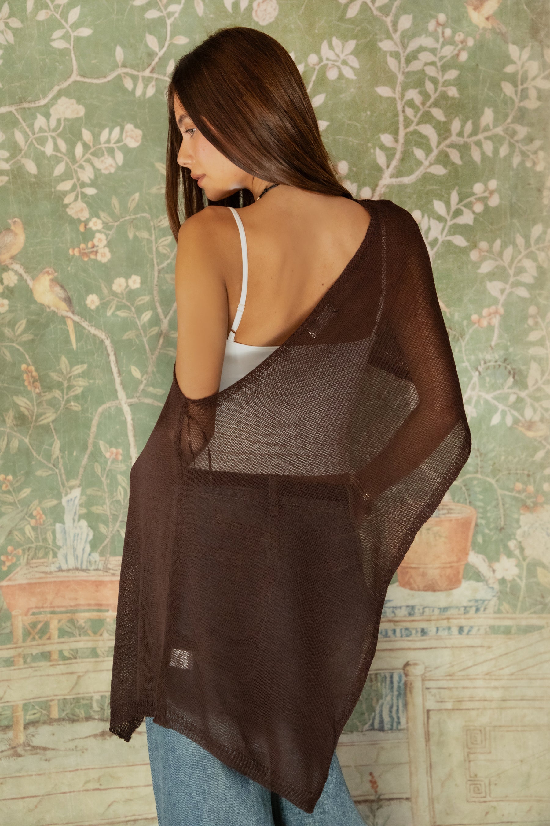 Woman wearing TIC TOC TC31873 SOLEI lightweight open sheer sweater knit poncho in BROWN, featuring a wide off-shoulder neckline, airy open knit texture, relaxed draped poncho silhouette, and asymmetrical hem, styled over a white cami with light wash denim for a western-casual summer layering look.